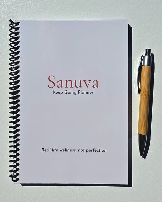 A spiral-bound planner titled 'Sanuva Keep Going Planner' lies on a light surface next to a bamboo pen.
