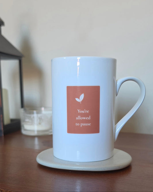 A white ceramic mug with a terracotta-colored square label that reads 'You're allowed to pause' with a small leaf icon, sitting on a beige coaster on a wooden table.