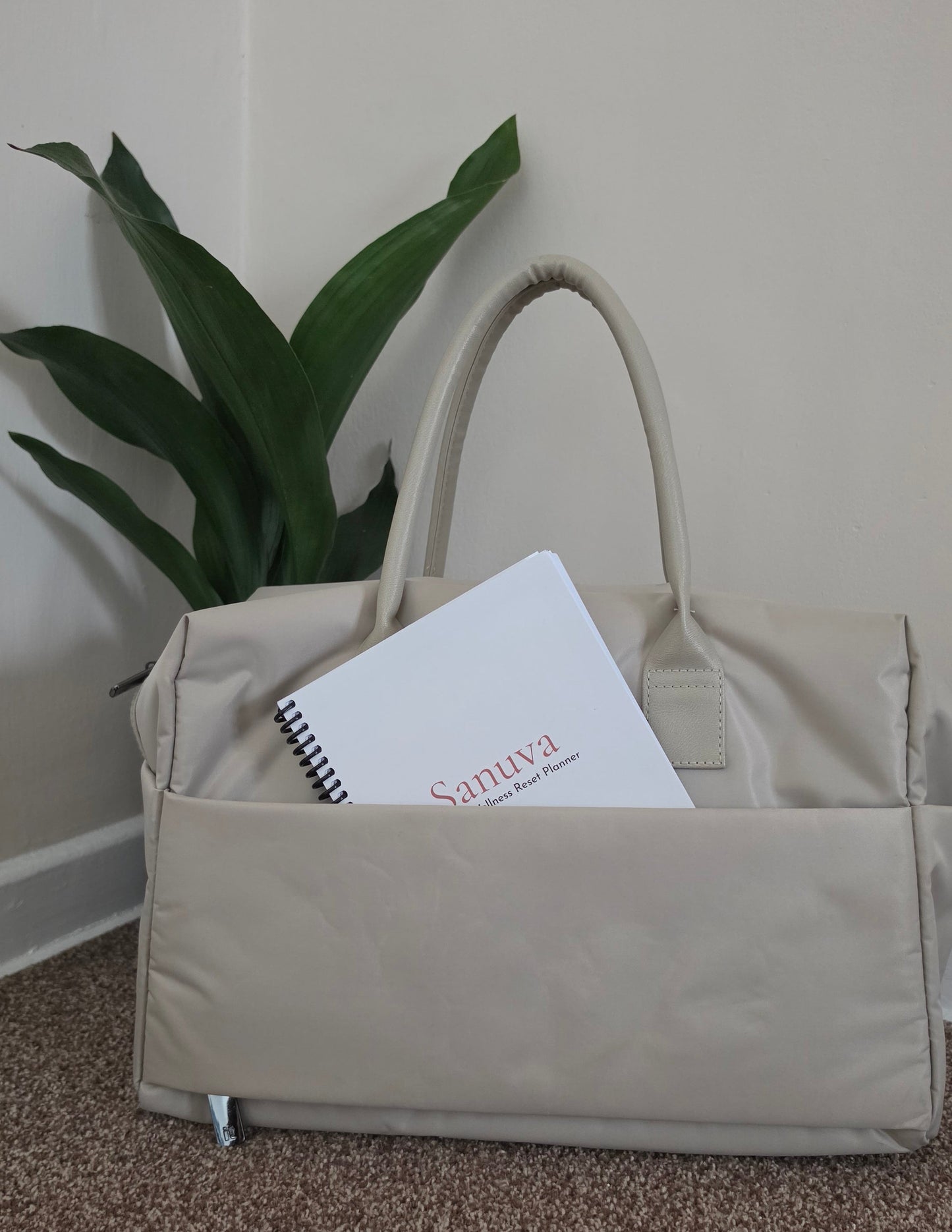 A beige bag with dual top handles sits on a carpeted floor against a plain wall, with a green houseplant behind it and a journal partially tucked into the front pocket.