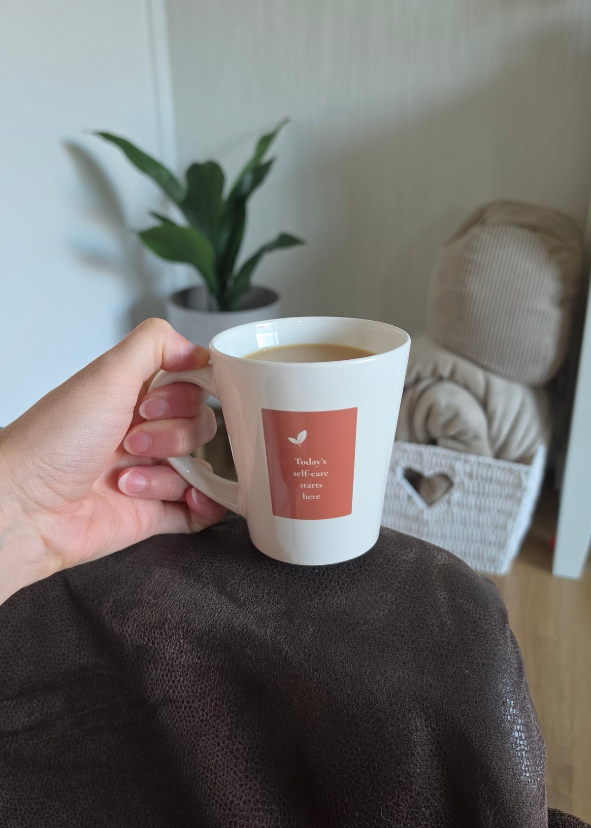 A hand holds a white ceramic mug with a terracotta-colored square label - filled with tea. The mug is positioned over a dark brown textured sofa arm in a cozy indoor setting with a potted plant and a basket in the background.