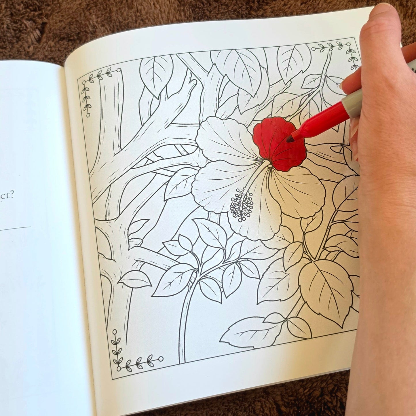 A hand holding a red marker coloring a black-and-white floral line drawing in an open coloring book, with a brown textured surface underneath.