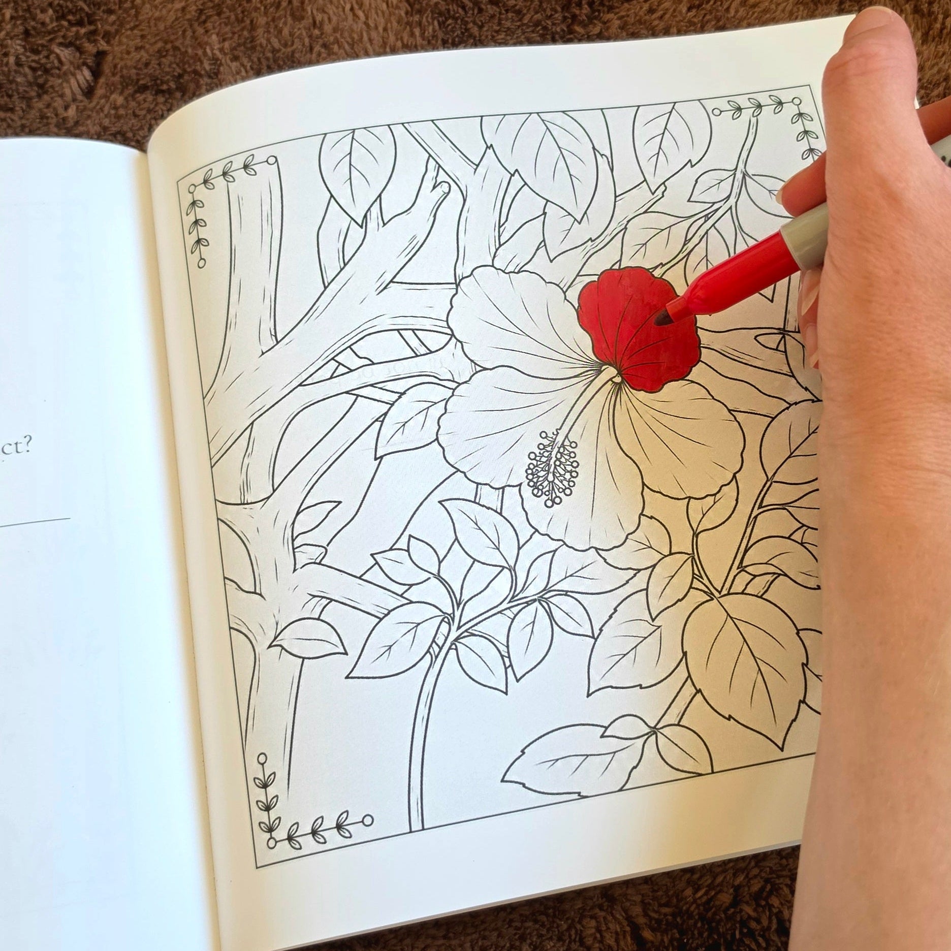 A hand holding a red marker coloring a black-and-white floral line drawing in an open coloring book, with a brown textured surface underneath.