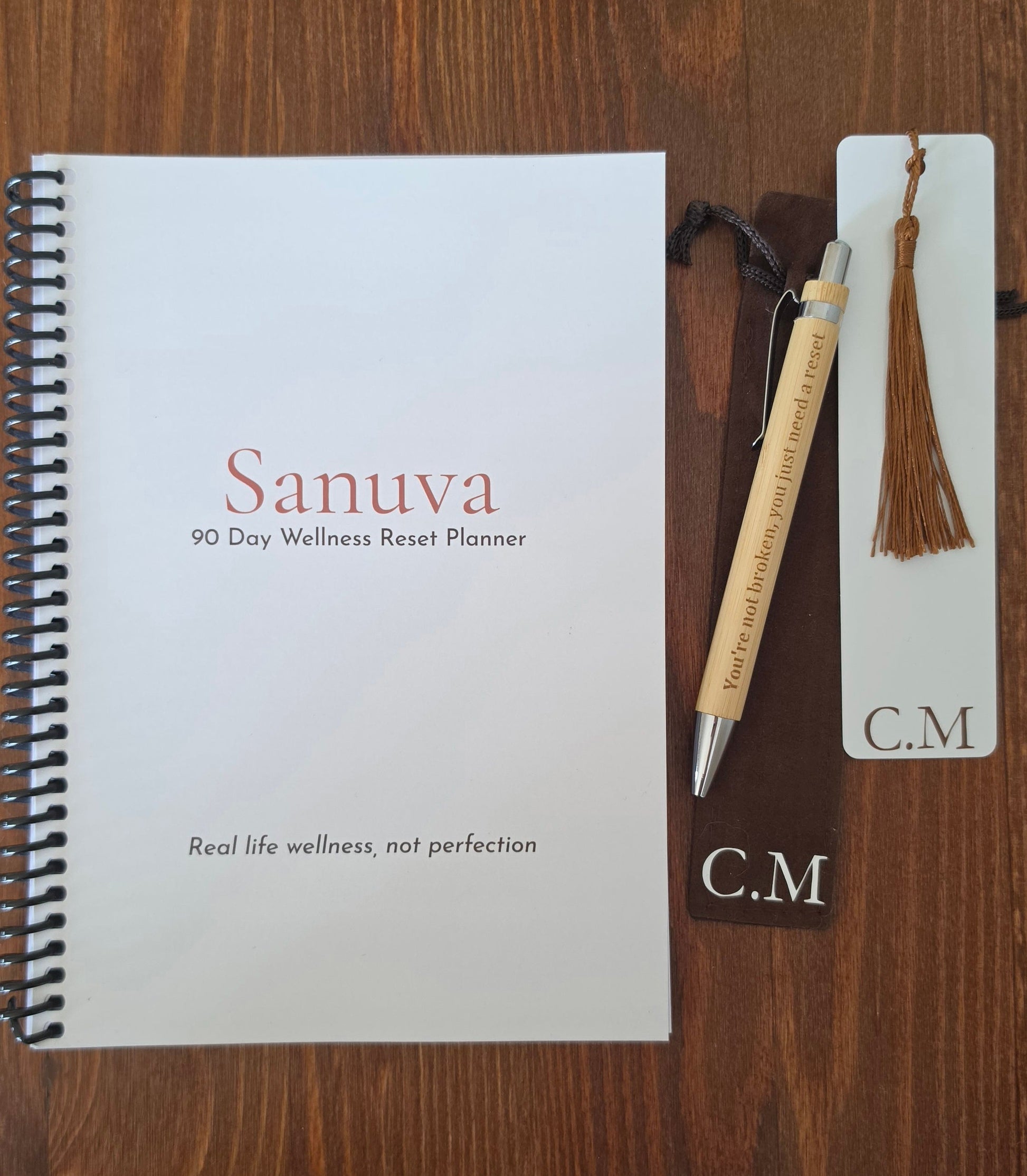 A spiral-bound 90 Day Wellness Reset Planner titled 'Sanuva' on a white cover, displayed on a dark wood surface alongside a wooden pen engraved with 'Real life wellness, not perfection' and a white tag with a brown tassel bookmark labeled 'C.M'.