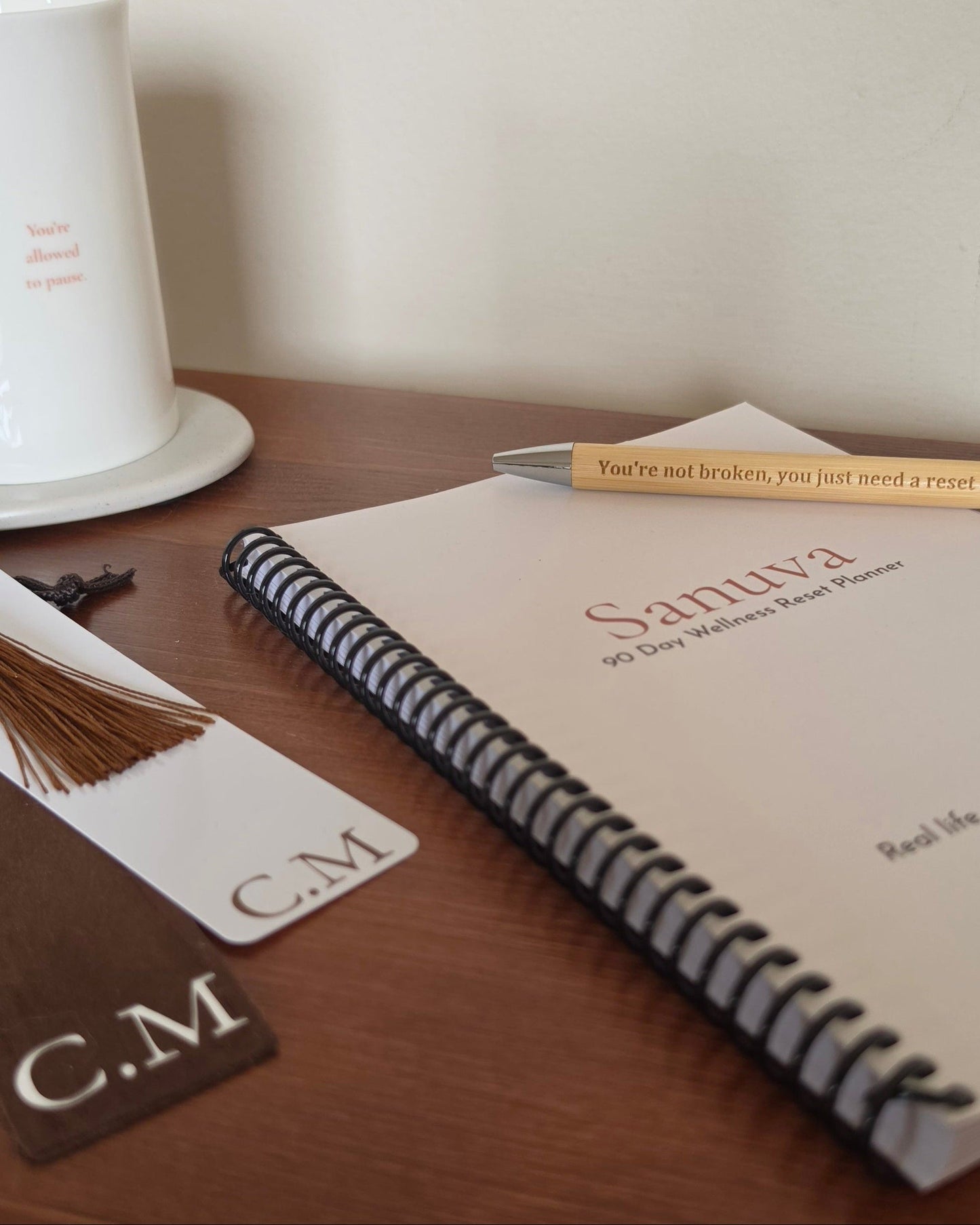 A spiral-bound planner or notebook on a wooden table with a pen resting across the cover. The cover reads 'Sanuva 90 Day Walker's Reset Planner.