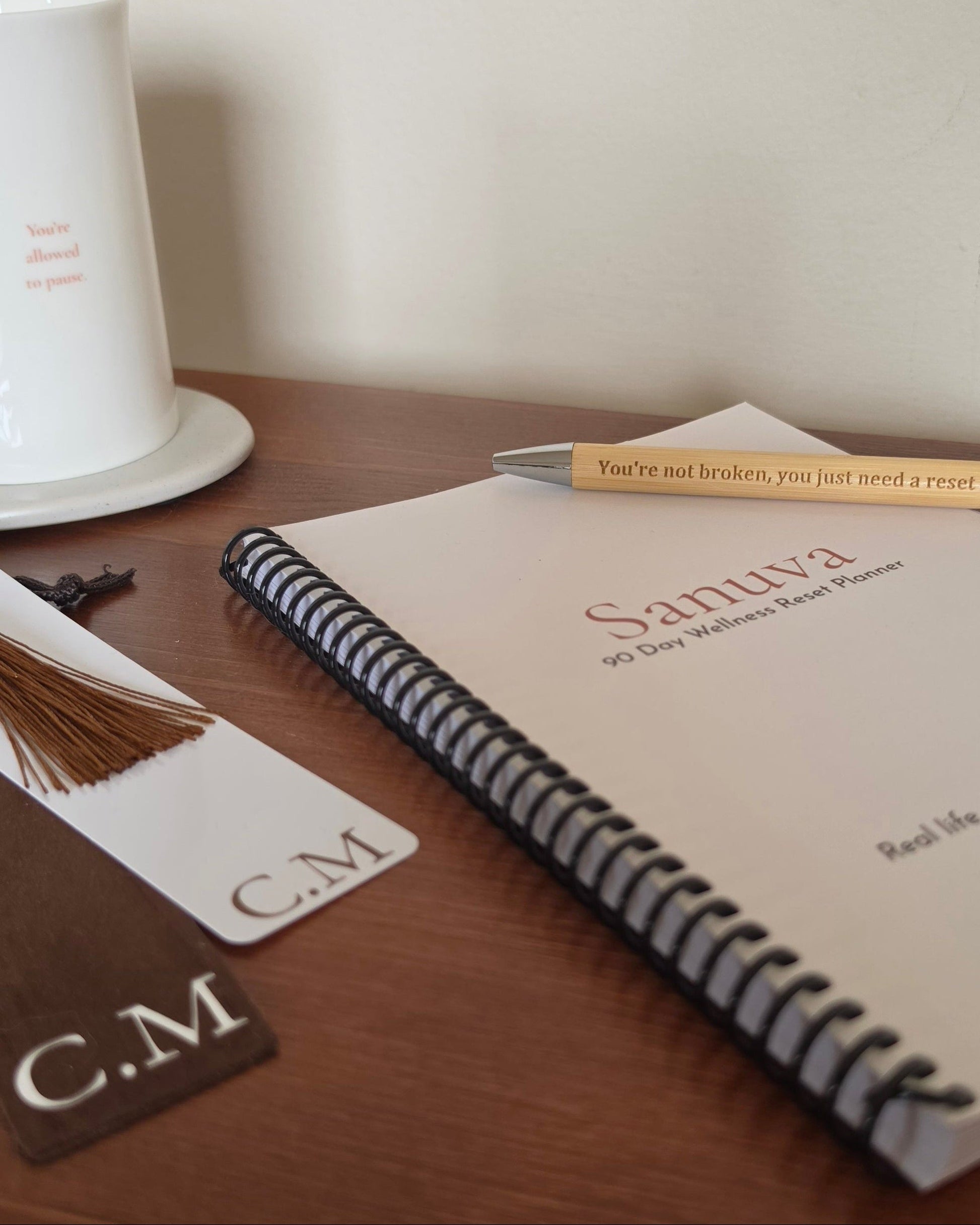 A spiral-bound planner or notebook on a wooden table with a pen resting across the cover. The cover reads 'Sanuva 90 Day Walker's Reset Planner.