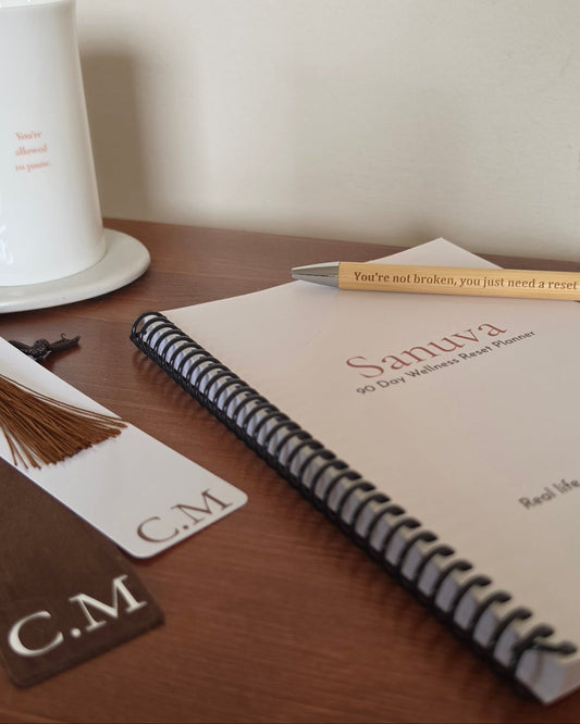 A spiral-bound planner or notebook on a wooden table with a pen resting across the cover. The cover reads 'Sanuva 90 Day Walker's Reset Planner.