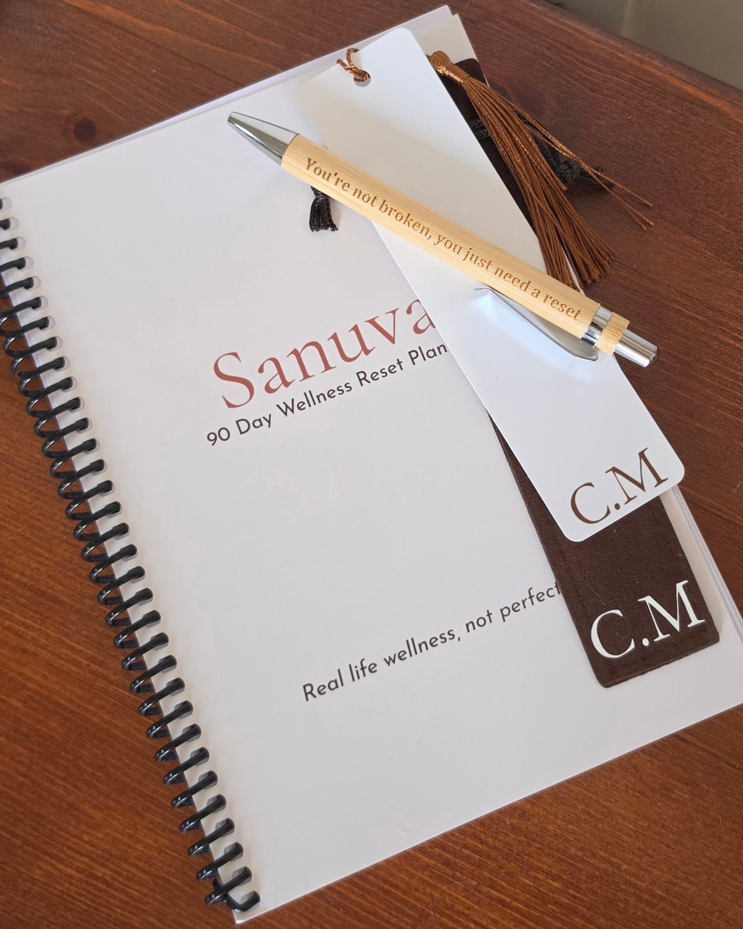 A spiral-bound notebook titled 'Sanuva 90 Day Wellness Reset Plan' lies on a wooden surface, with a wooden pen resting across the top and a branded tag attached. The pen is light wood with dark brown engraved text.