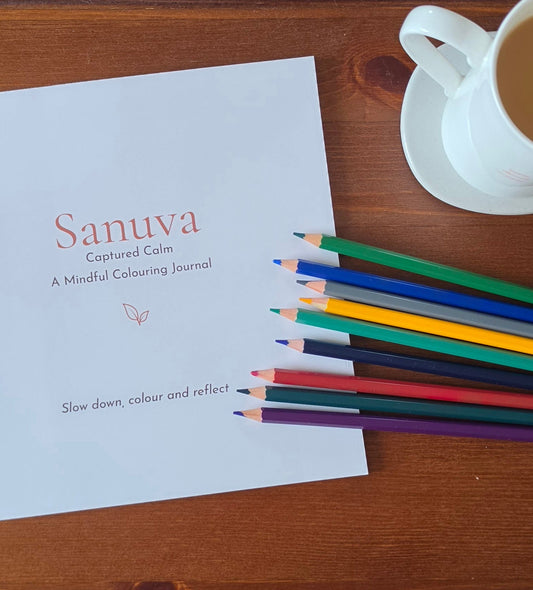 A white journal cover printed with the title 'Sanuva: Captured Calm' lies on a wooden table, with a row of colored pencils fanned across the lower right and a white coffee cup in the upper right corner.