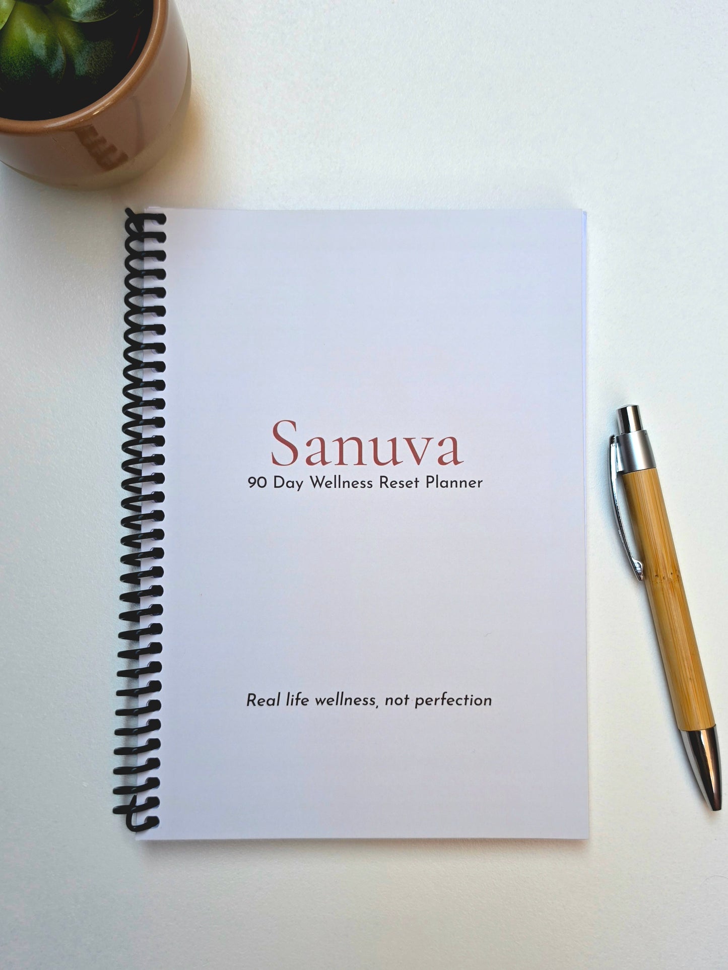 Sanuva 90 Day Wellness Reset Planner on a white surface with a pen and plant.