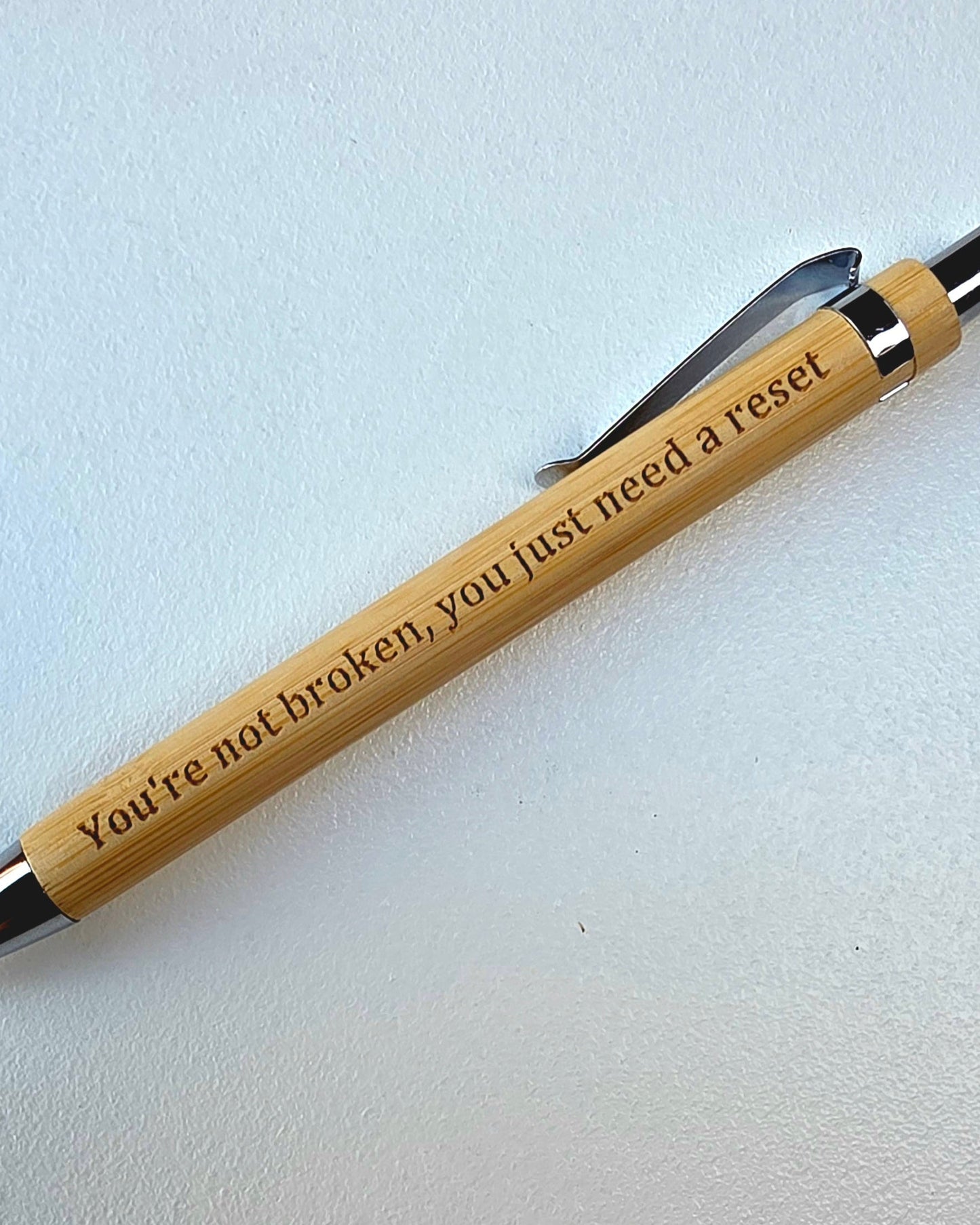 A bamboo pen with engraved text reading 'You're not broken, you just need a reset' and silver metal accents, resting on a light gray textured surface.