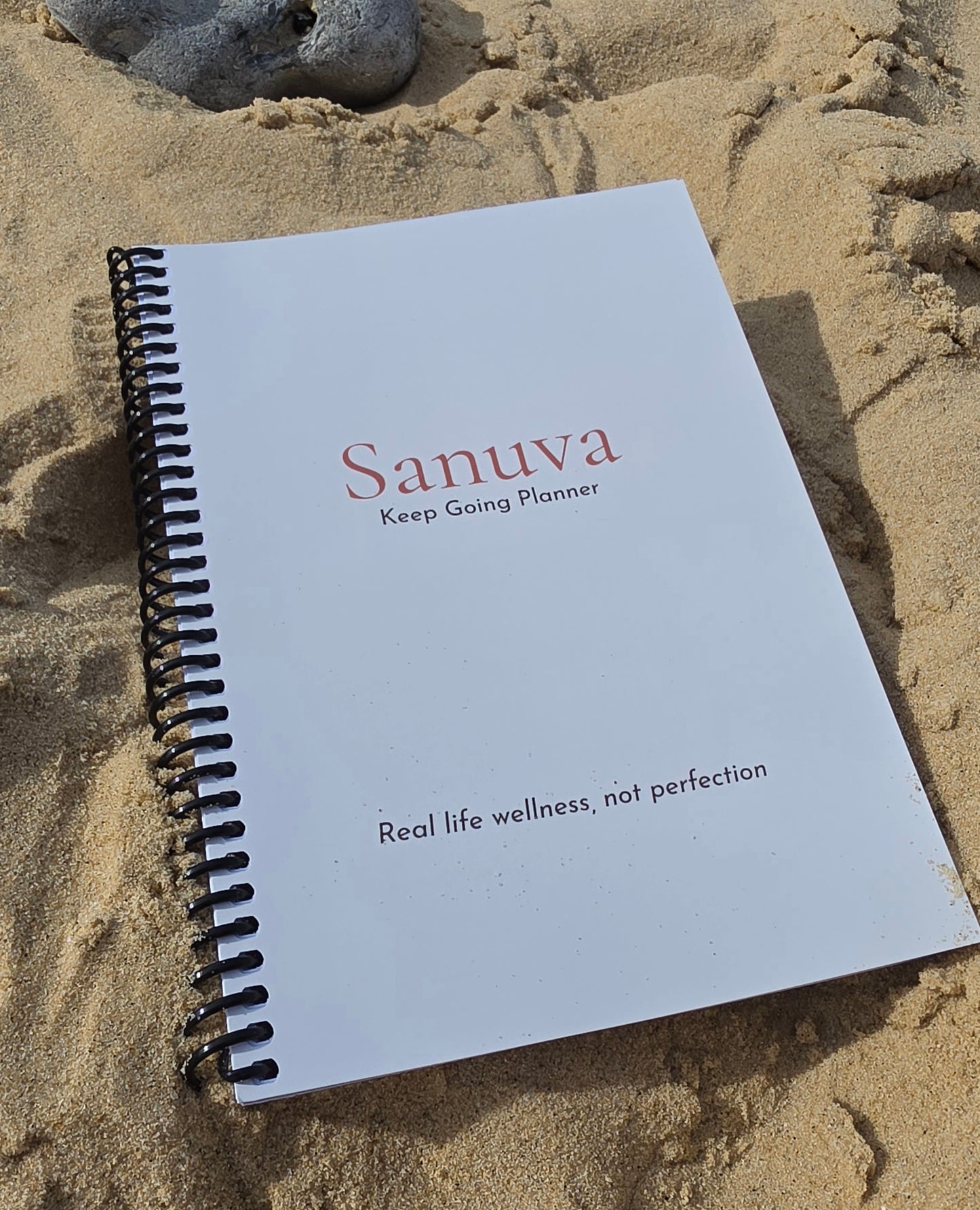 A spiral-bound notebook titled 'Sanuva Keep Going Planner' resting on sand.