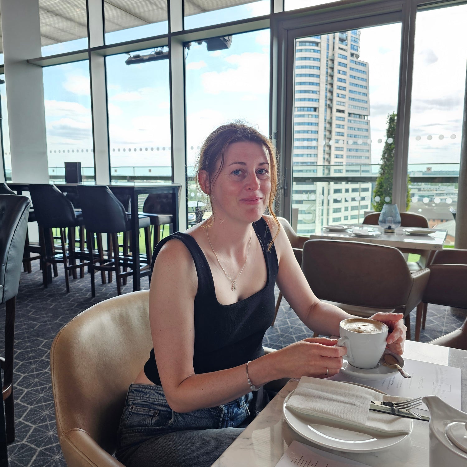A woman wearing a black tank top and denim bottoms sits at a cafe table holding a white mug, with a city skyline visible through large windows behind her.