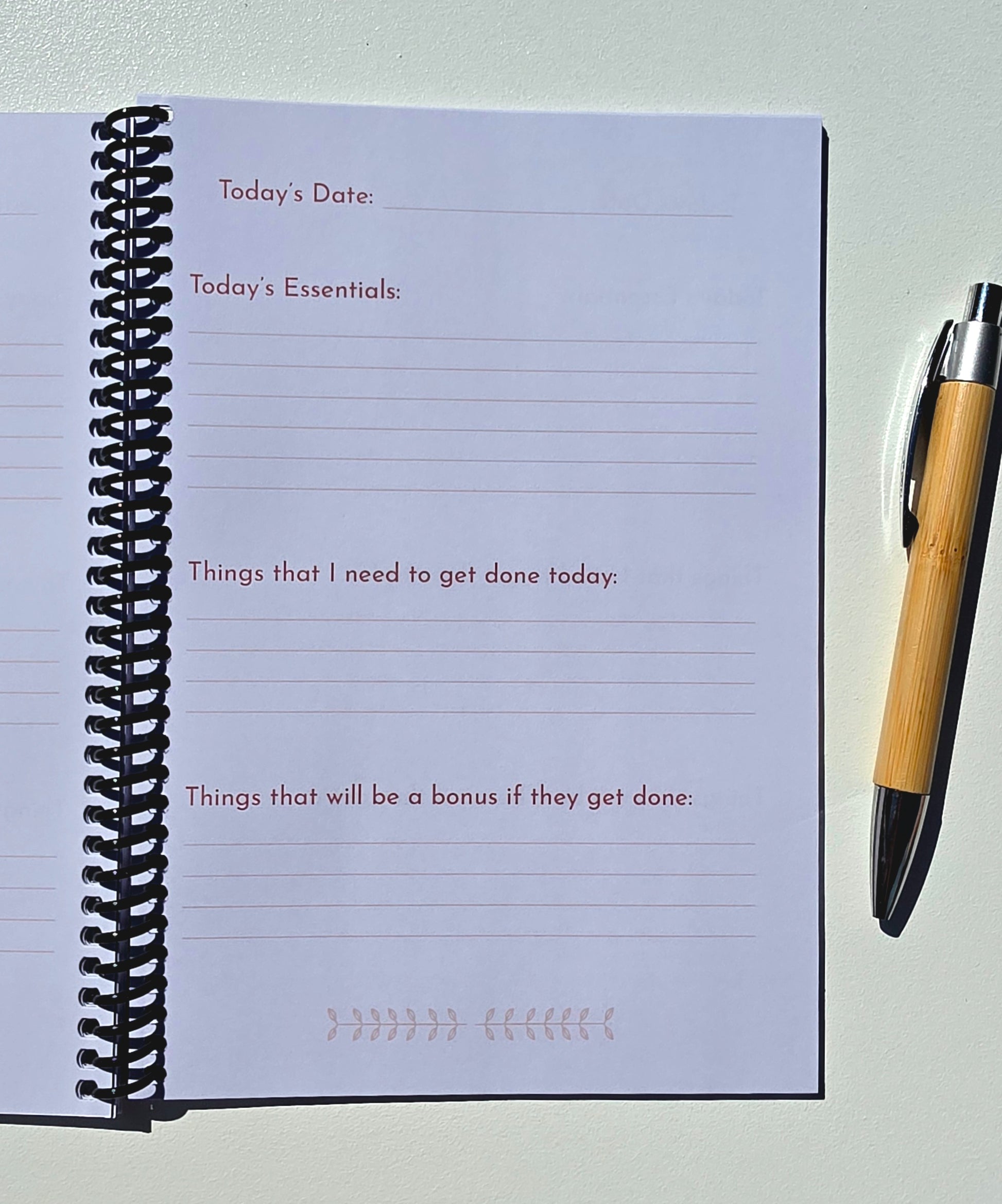 An open spiral-bound planner or notepad with lined writing pages, showing fields for today's date, today's essentials, and tasks, photographed on a light surface with a wooden pen beside it.