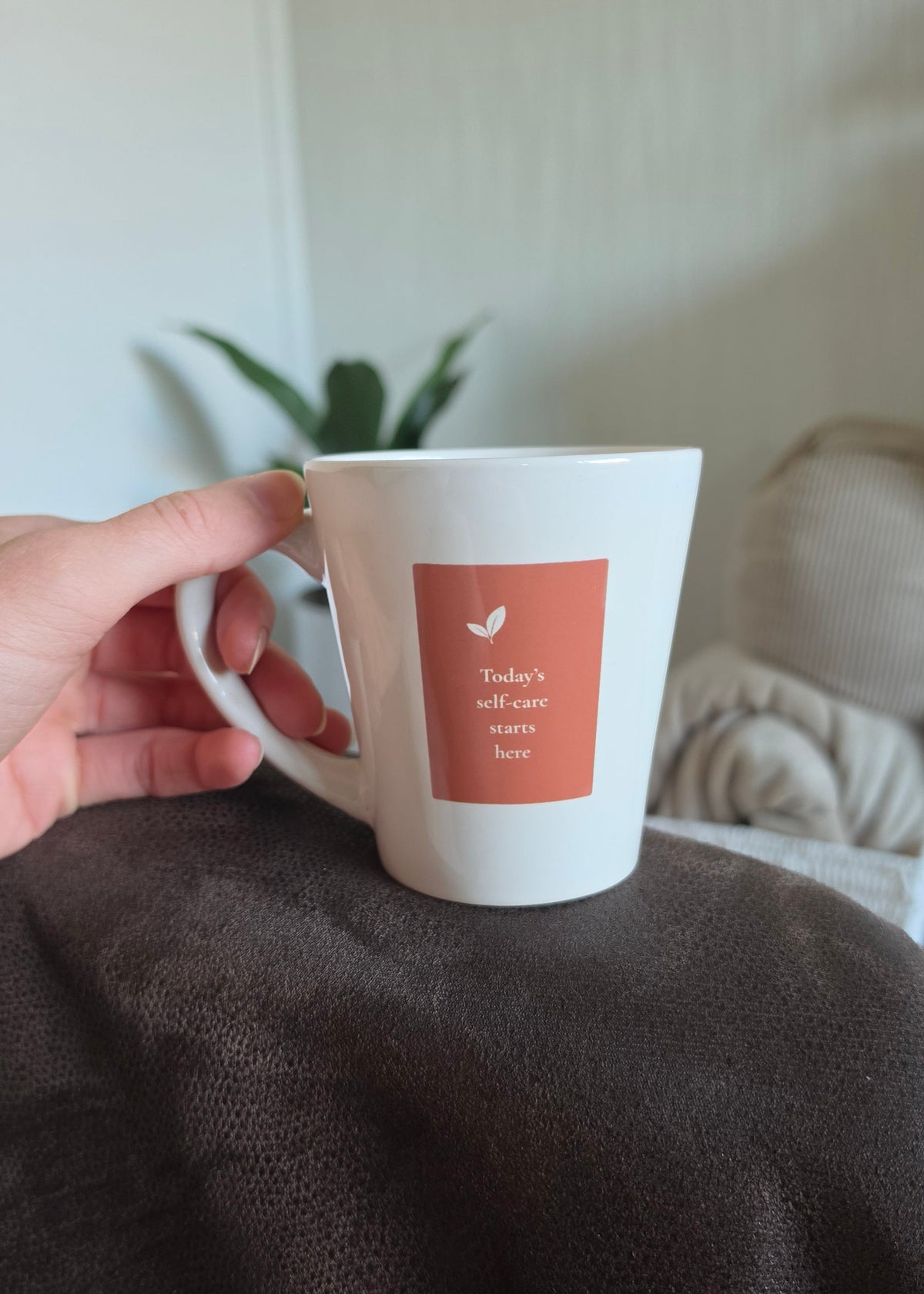A hand holding a white ceramic mug with a terracota square label that reads 'Today's self-care starts here' The mug is resting on a dark brown textured chair arm in a cozy indoor setting with a blurred plant and rolled blankets in the background.