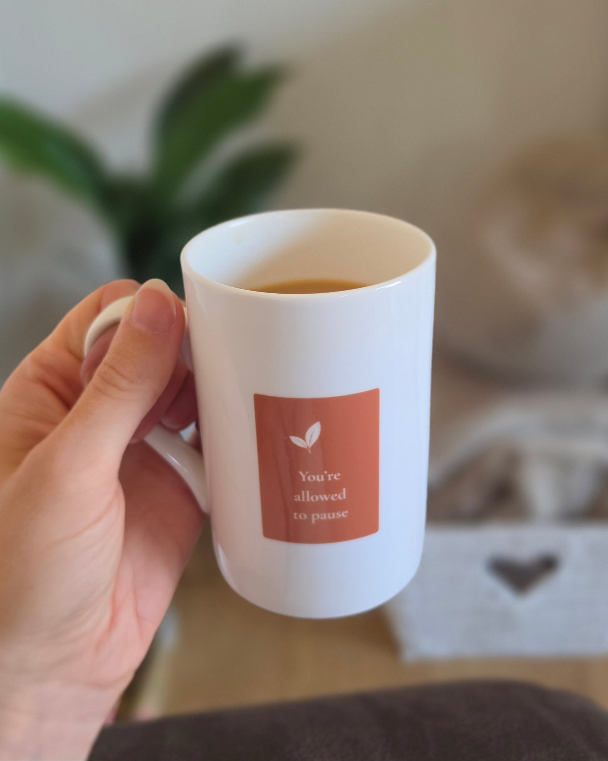A hand holding a white ceramic mug filled with tea, featuring a terracotta square label with a small leaf icon and the text 'You're allowed to pause'. The background is a softly blurred indoor setting with a plant and neutral decor.