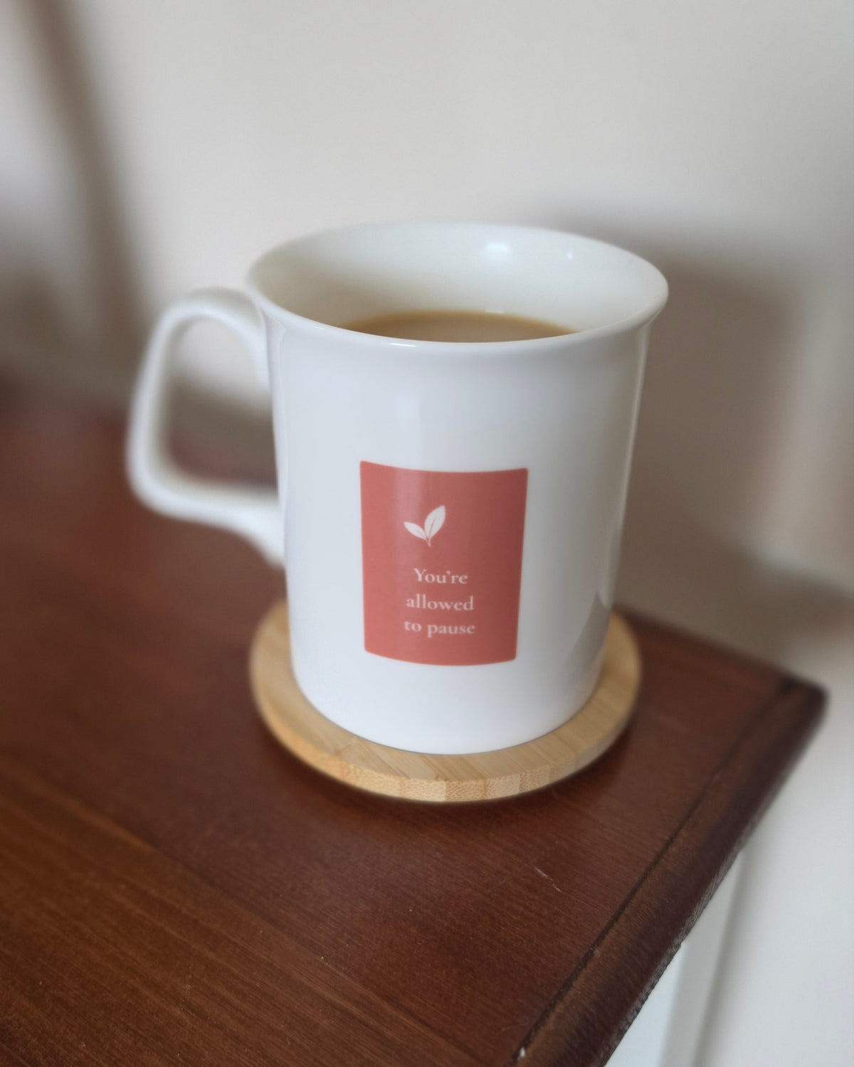 A white ceramic mug filled with tea sits on a round wooden coaster on a dark wooden table. The mug features a square label with 'You're allowed so pause'.