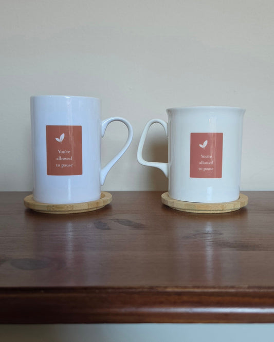 Two white ceramic mugs with light wood coasters sit on a dark wooden table against a plain beige wall. Each mug has a square terracotta-colored label featuring a small leaf and the text 'You're allowed to pause'.