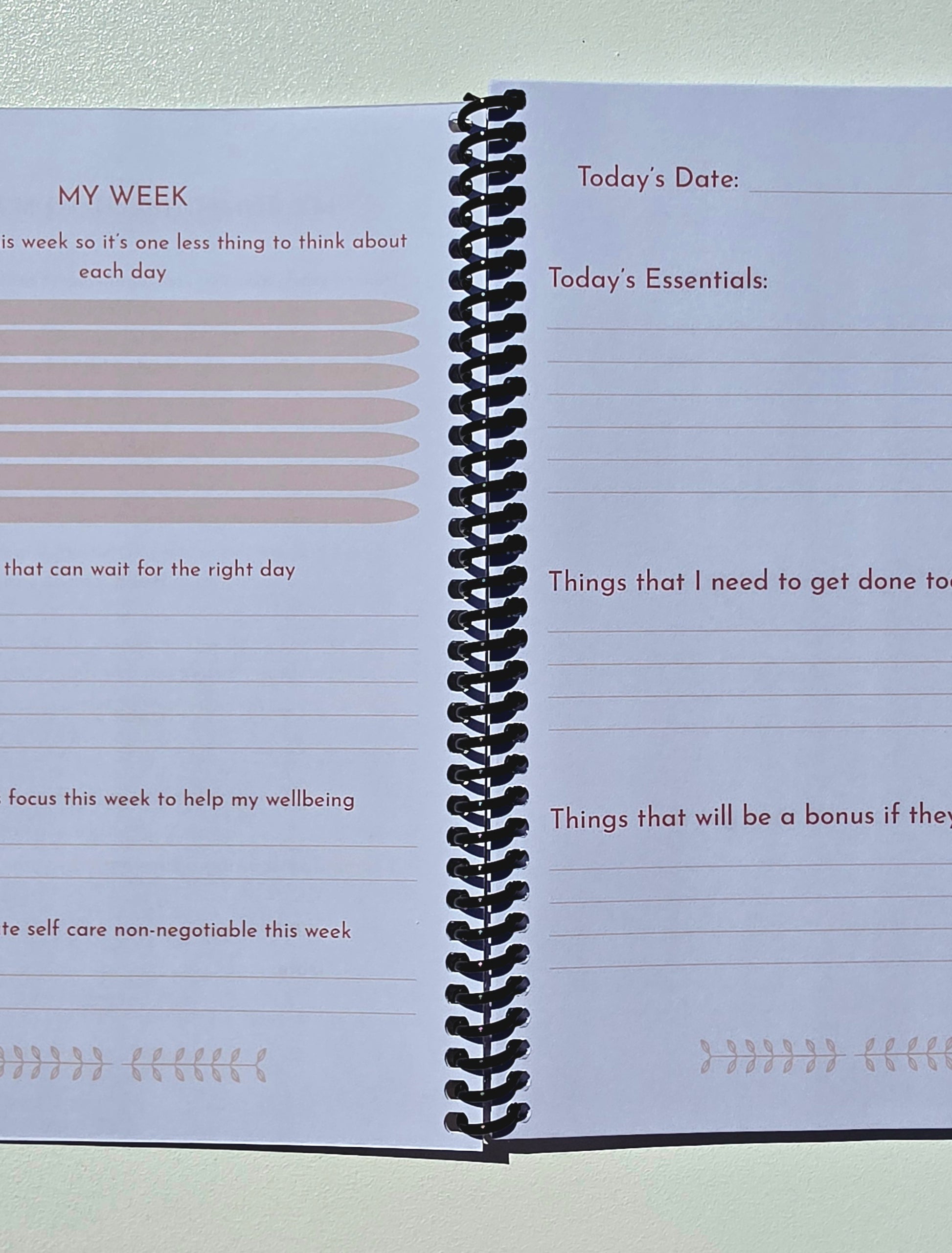 An open spiral-bound planner or notebook showing two lined pages with prompts such as 'My Week,' 'Today's Date,' and 'Today's Essentials.' The pages are white with light beige lines and dark brown text, bound by a black spiral.