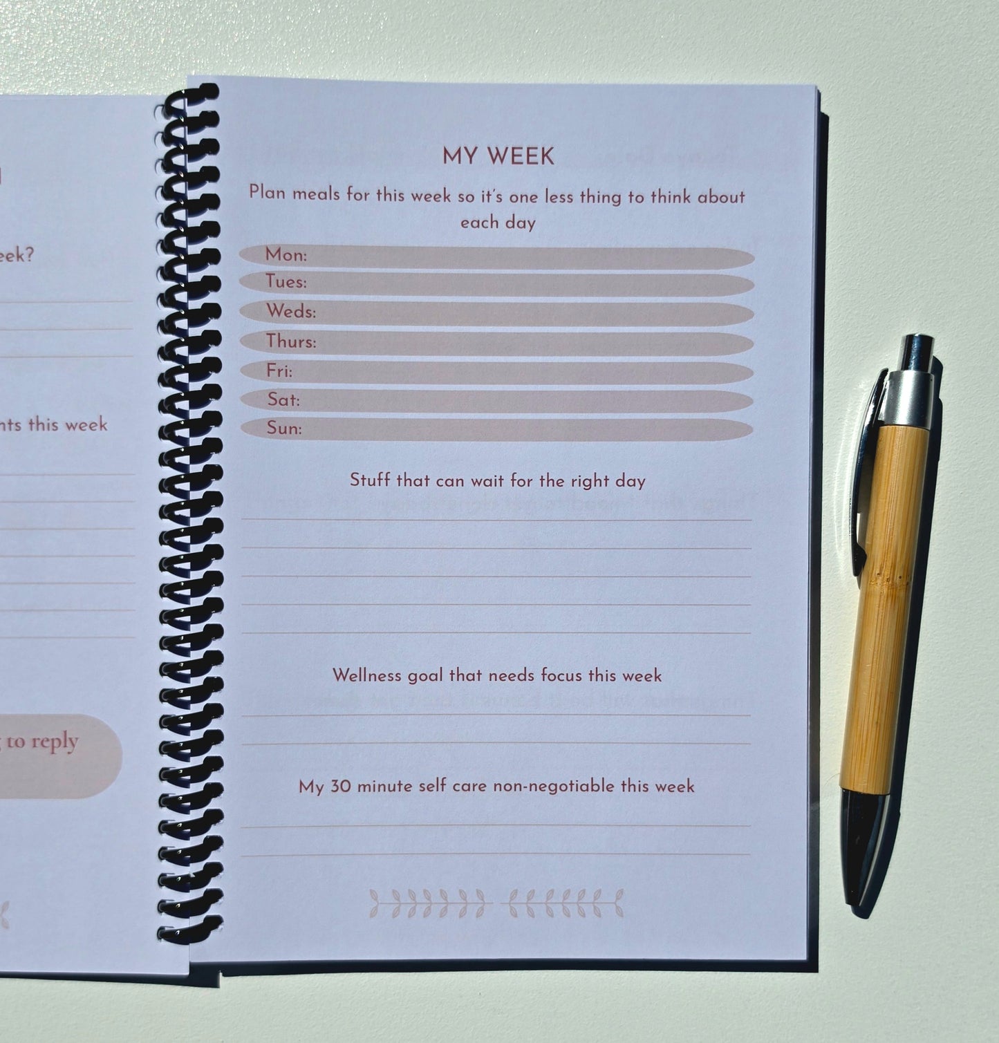 An open spiral-bound planner or journal showing a weekly reflection page with fields to plan meals, list days of the week, and write prompts, photographed next to a bamboo pen on a light background.