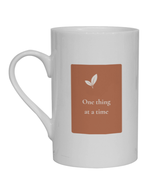 A white ceramic mug with a terracota rectangular label on the front featuring a small leaf graphic and the text 'One thing at a time' in white, shown on a plain white background.