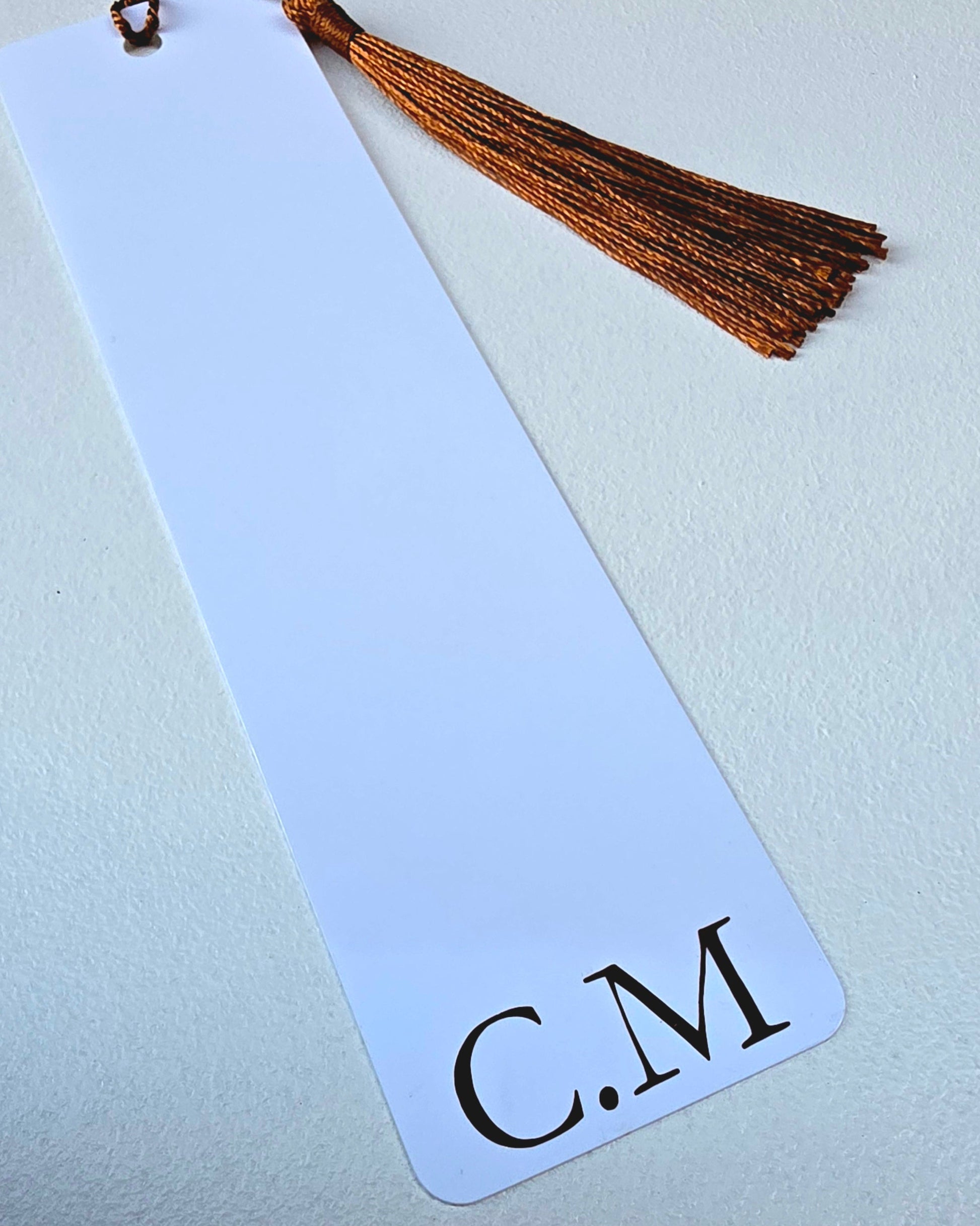 A white rectangular bookmark with rounded corners, a brown tassel at the top, and the black initials 'C.M' printed at the bottom, shown on a light textured background.