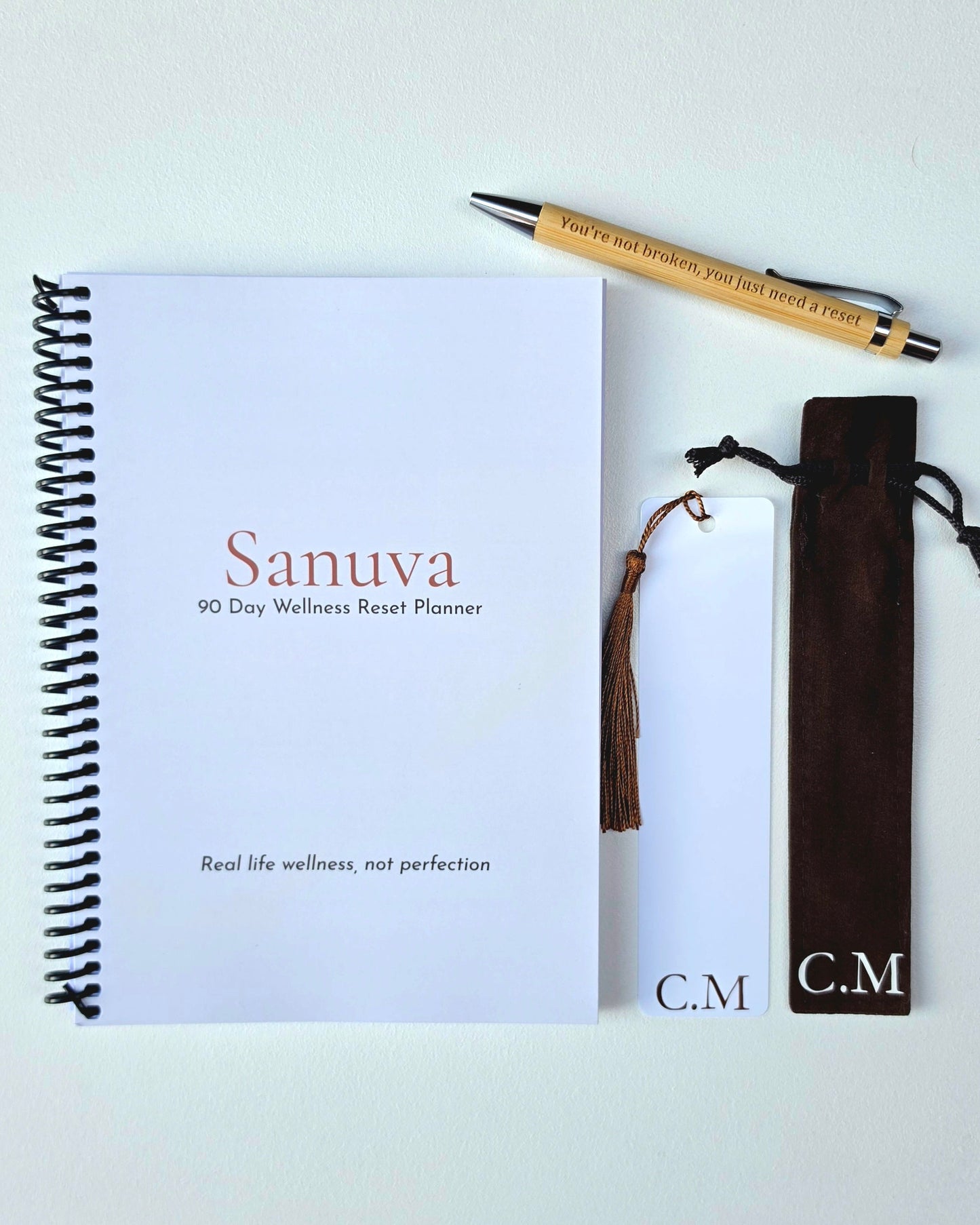 A flat-lay of a spiral-bound planner titled 'Sanuva 90 Day Wellness Reset Planner' on a white background, accompanied by a wooden pen, a brown tassel bookmark, and a dark brown velvet pouch with 'C.M' monogrammed on the front.