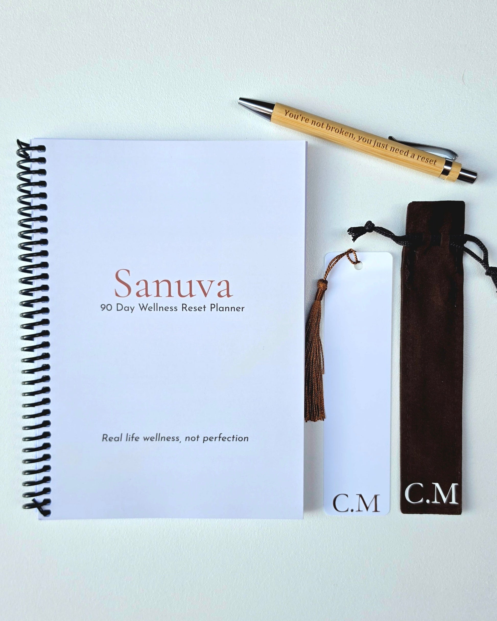 A flat-lay of a spiral-bound planner titled 'Sanuva 90 Day Wellness Reset Planner' on a white background, accompanied by a wooden pen, a brown tassel bookmark, and a dark brown velvet pouch with 'C.M' monogrammed on the front.