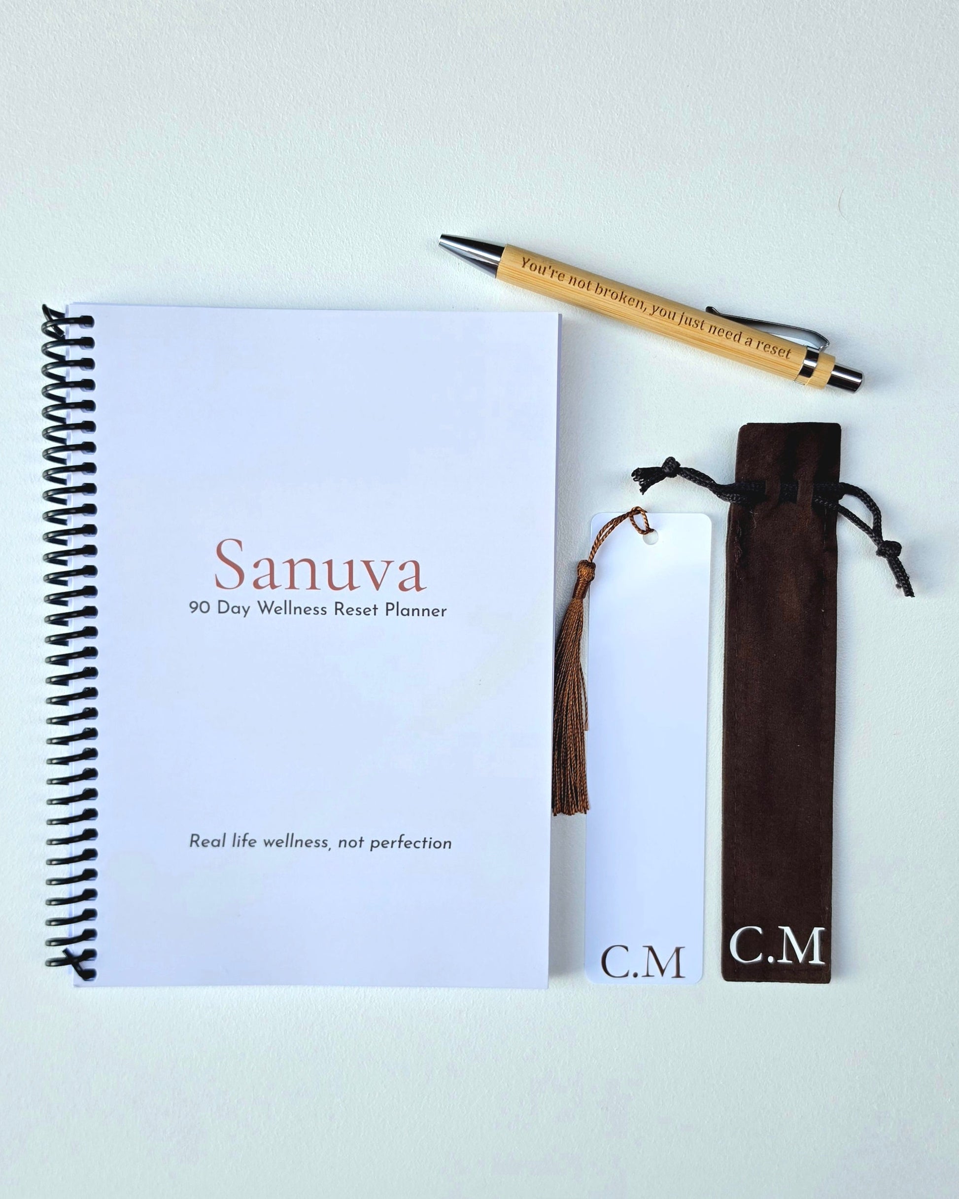 A spiral-bound planner notebook labeled 'Sanuva 90 Day Wellness Reset Planner' on a white cover, arranged with a bamboo pen, a white bookmark, and a dark brown tassel pouch on a light background.