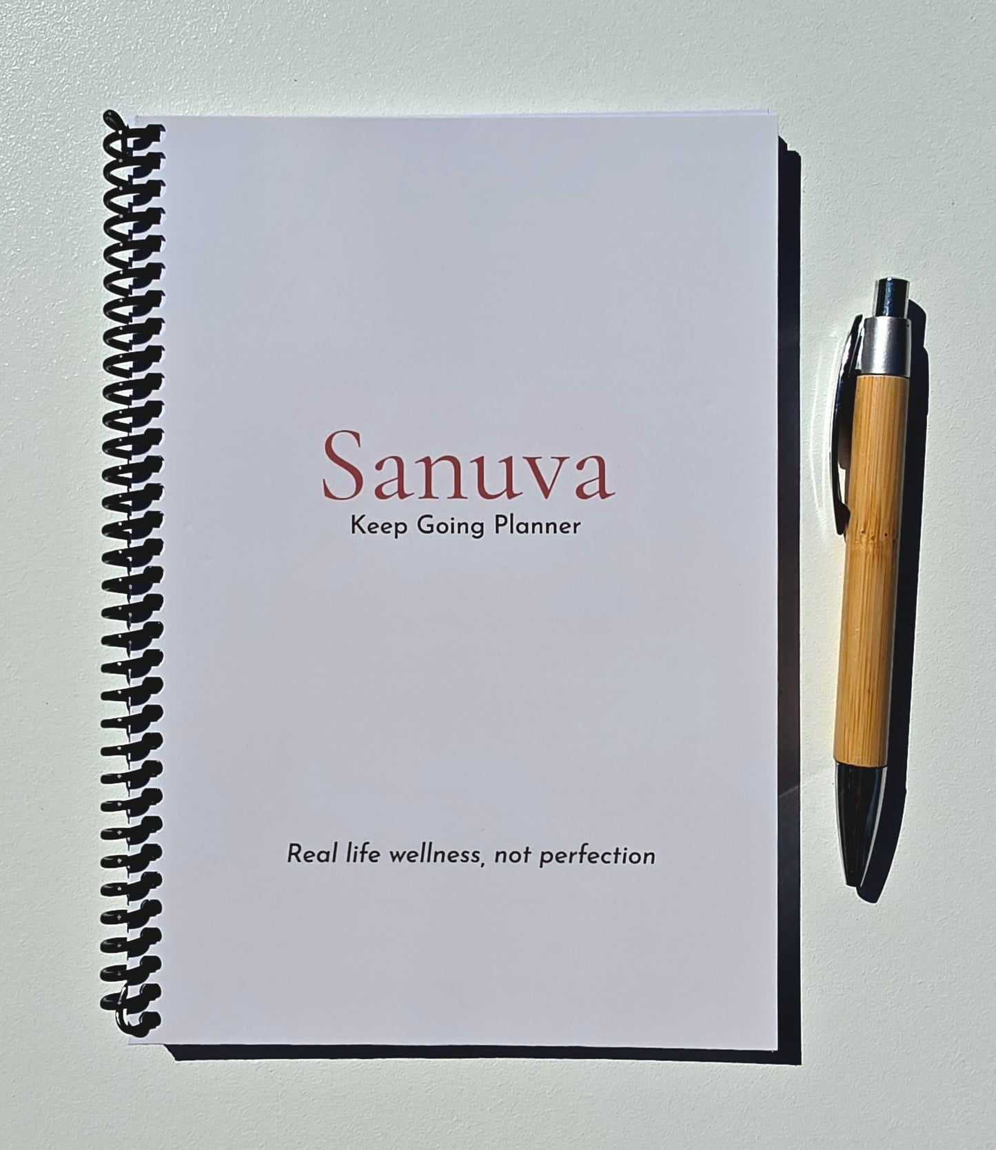 A spiral-bound planner titled 'Sanuva Keep Going Planner' lies on a light surface next to a bamboo pen.
