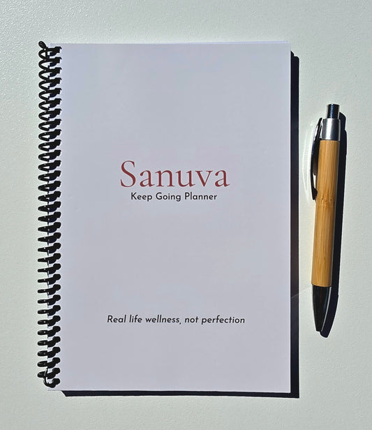 A spiral-bound planner titled 'Sanuva Keep Going Planner' lies on a light surface next to a bamboo pen.