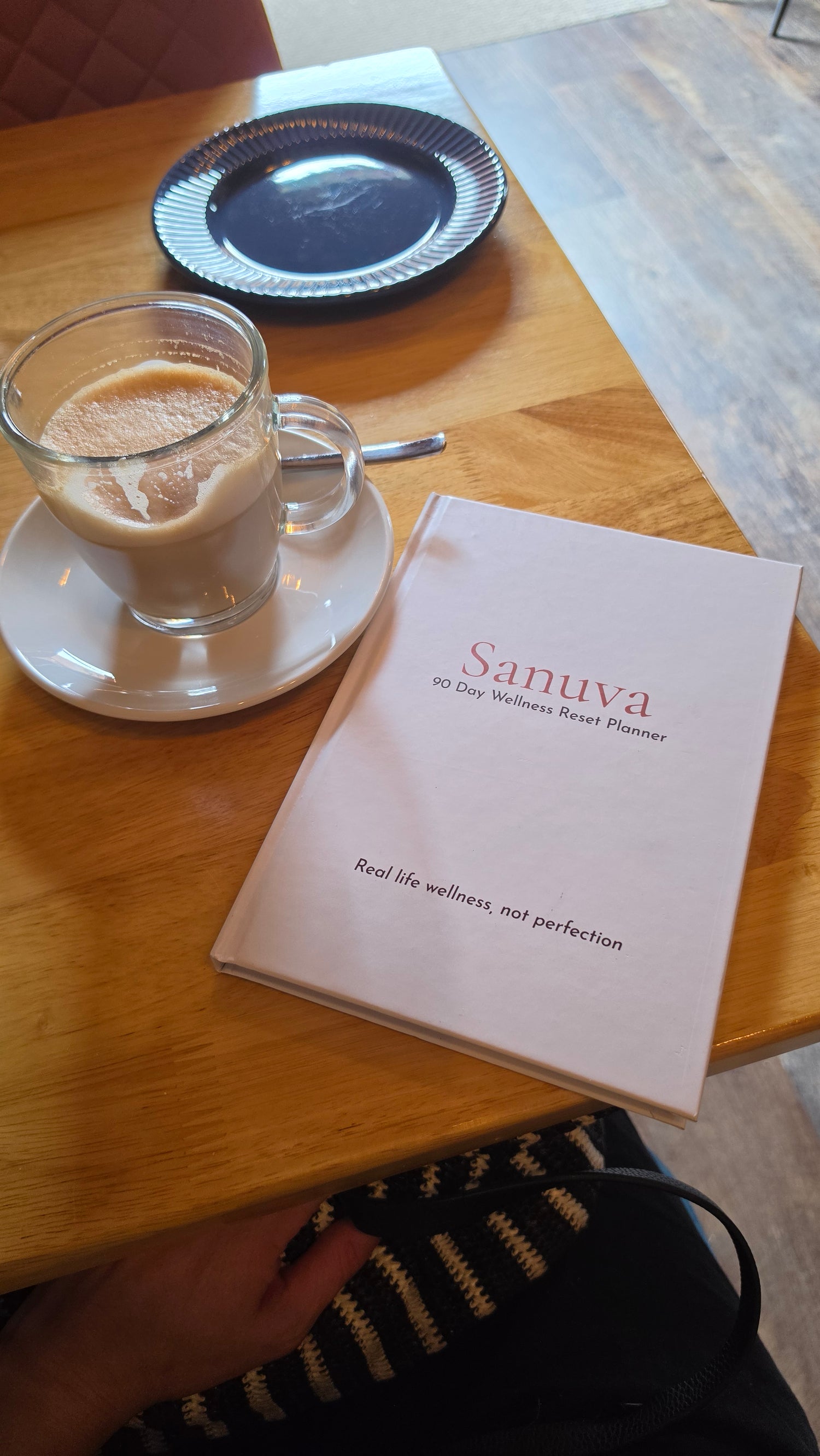 A close-up of a wooden table with a book printed with the brand name 'Sanuva', alongside a glass of coffee on a saucer.