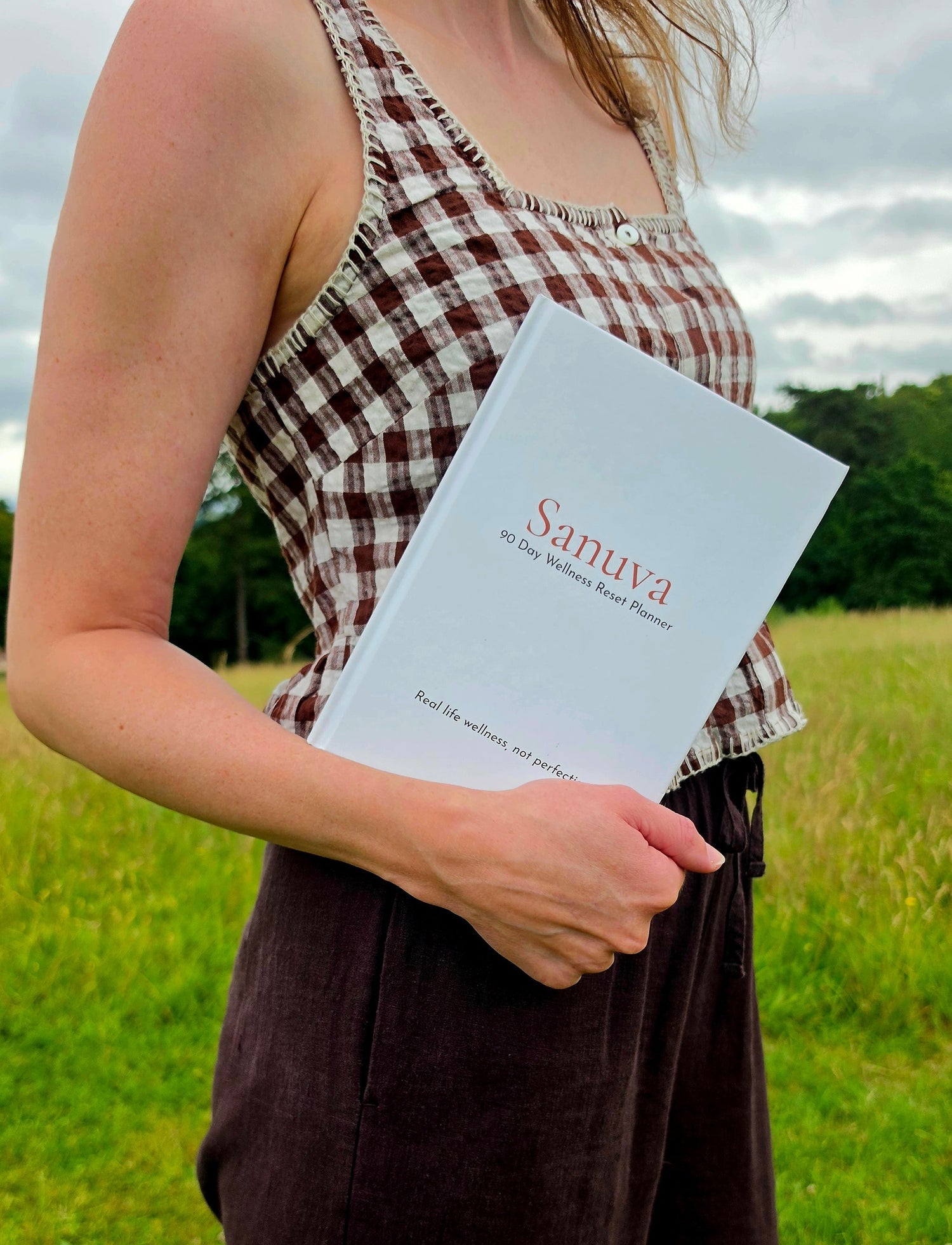 A woman standing in a grassy field holds a white book titled 'Sanuva' She is wearing a brown and white checkered sleeveless top and dark brown pants.