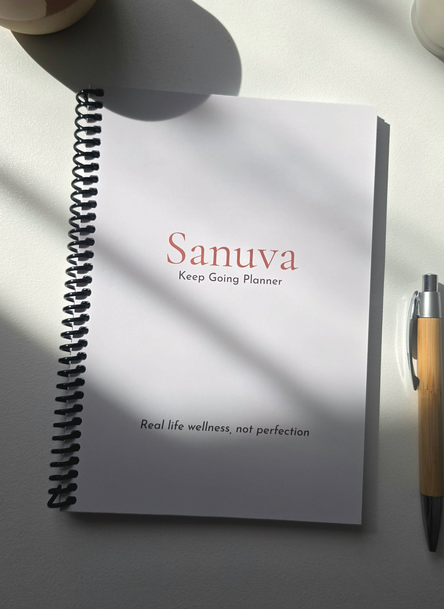 A spiral-bound planner notebook titled 'Sanuva Keep Going Planner' on a light surface with dramatic diagonal sunlight shadows, alongside a wooden pen.