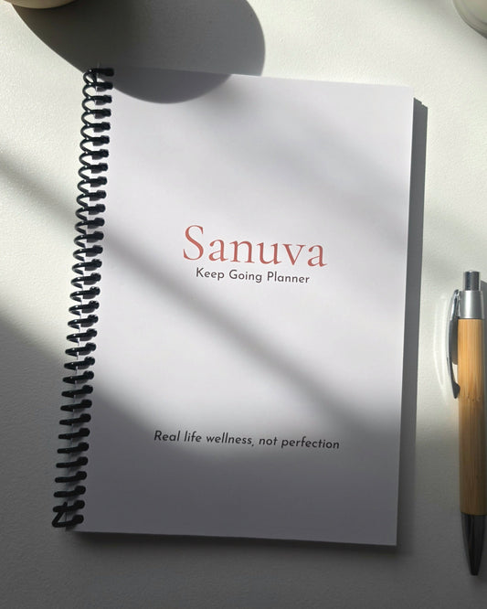 A spiral-bound planner notebook titled 'Sanuva Keep Going Planner' on a light surface with dramatic diagonal sunlight shadows, alongside a wooden pen.