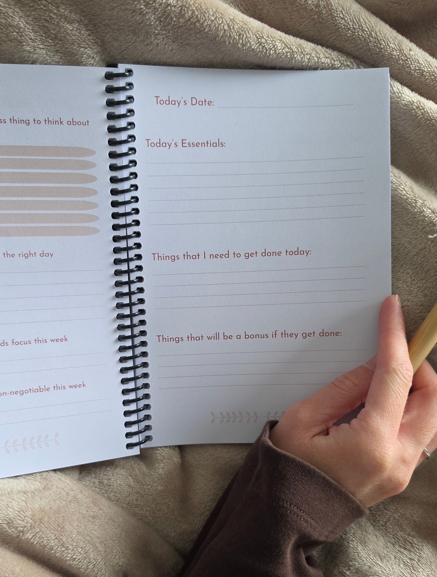 An open spiral-bound planner or journal lying on a beige blanket, with a hand holding a wooden pencil near the page. The page has lined writing space and prompts such as 'Today's Date' and 'Today's Essentials'.