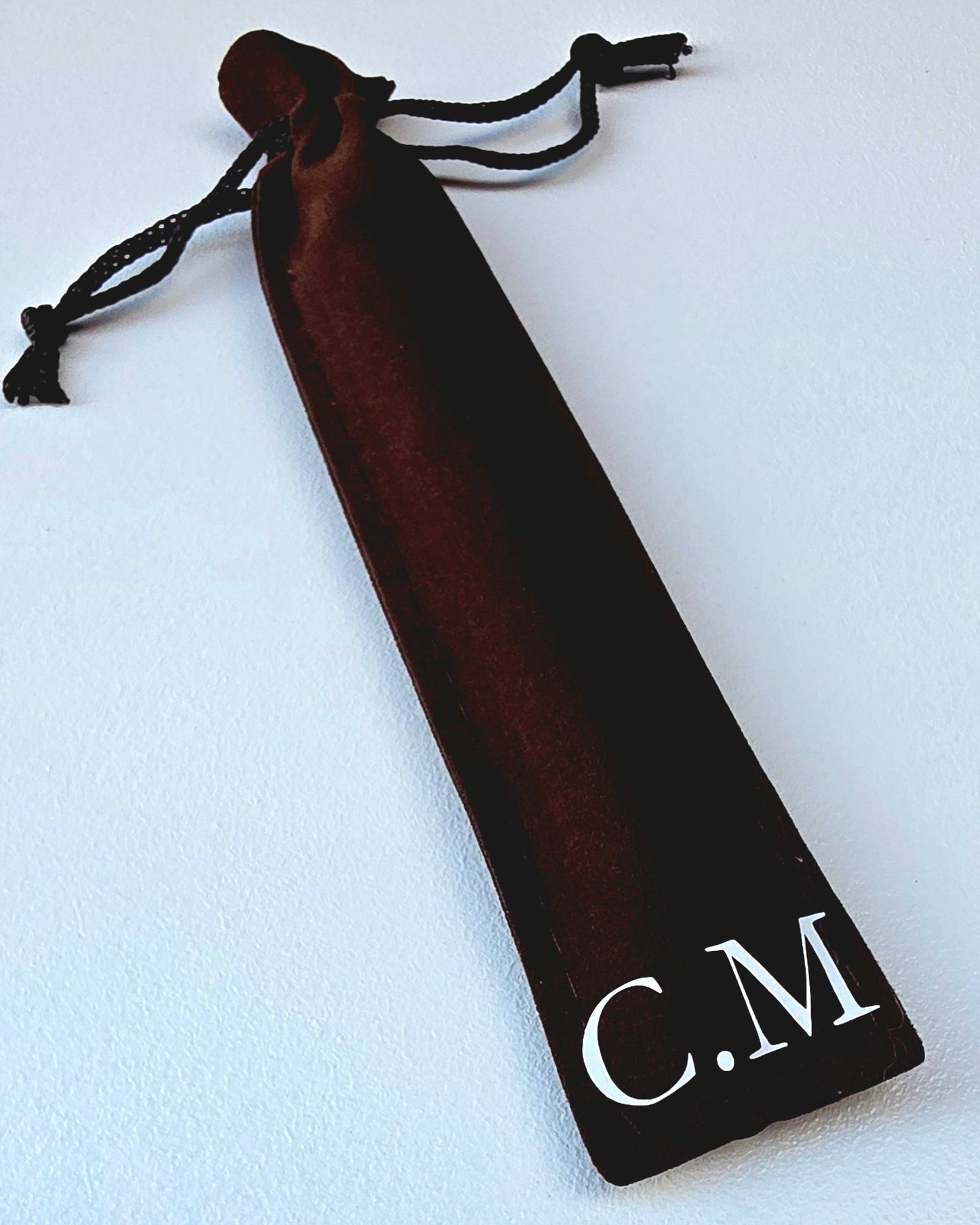 A dark brown velvet drawstring pouch with white monogram letters 'C.M' printed on the bottom corner, shown on a light textured background.