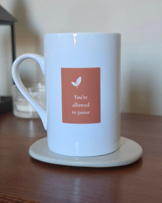 A white ceramic mug with a terracotta-colored square label that reads 'You're allowed to pause' with a small leaf icon, sitting on a light beige coaster on a wooden table.