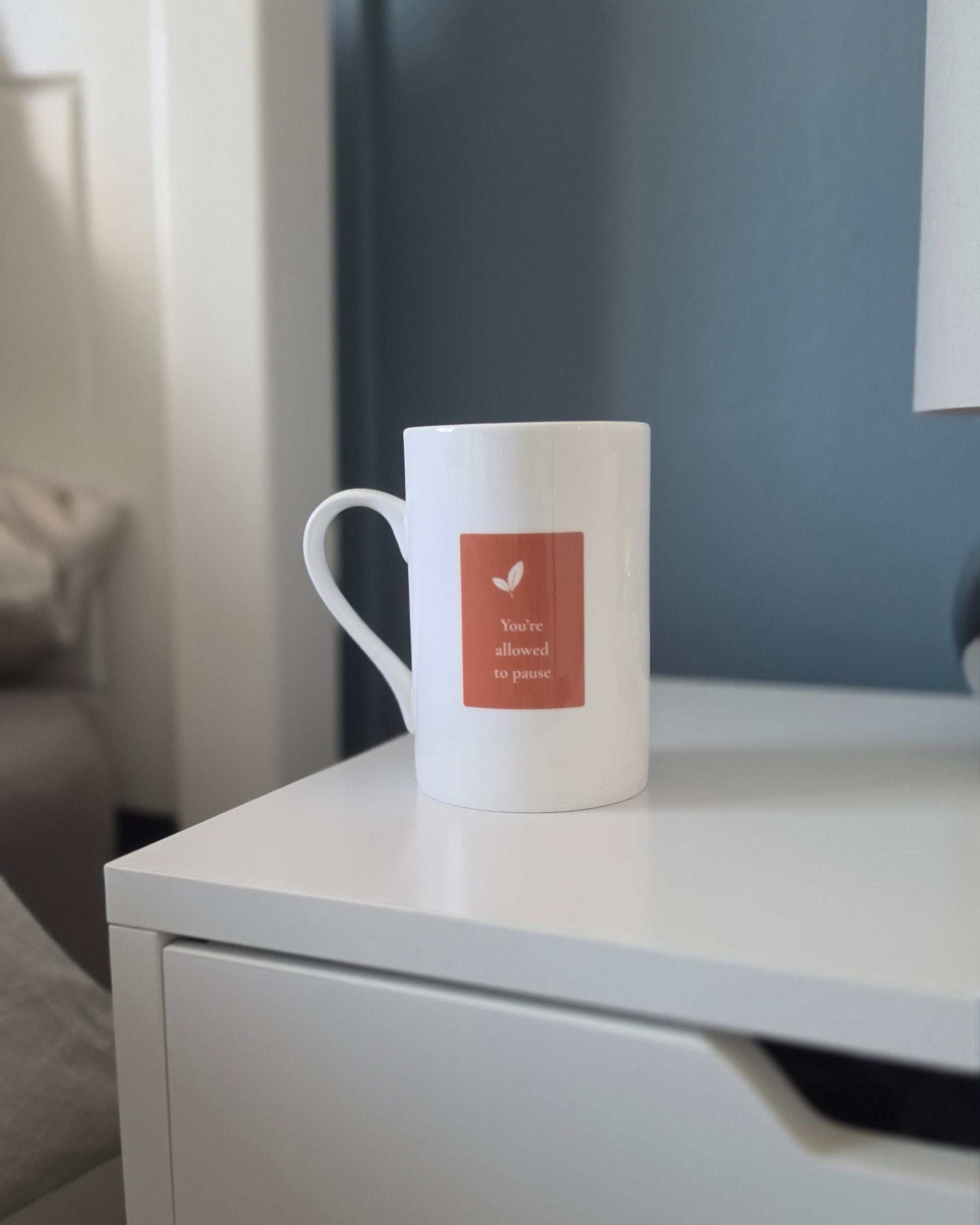 A white ceramic coffee mug with a terracotta square graphic on the front, placed on a white nightstand in a bedroom setting.