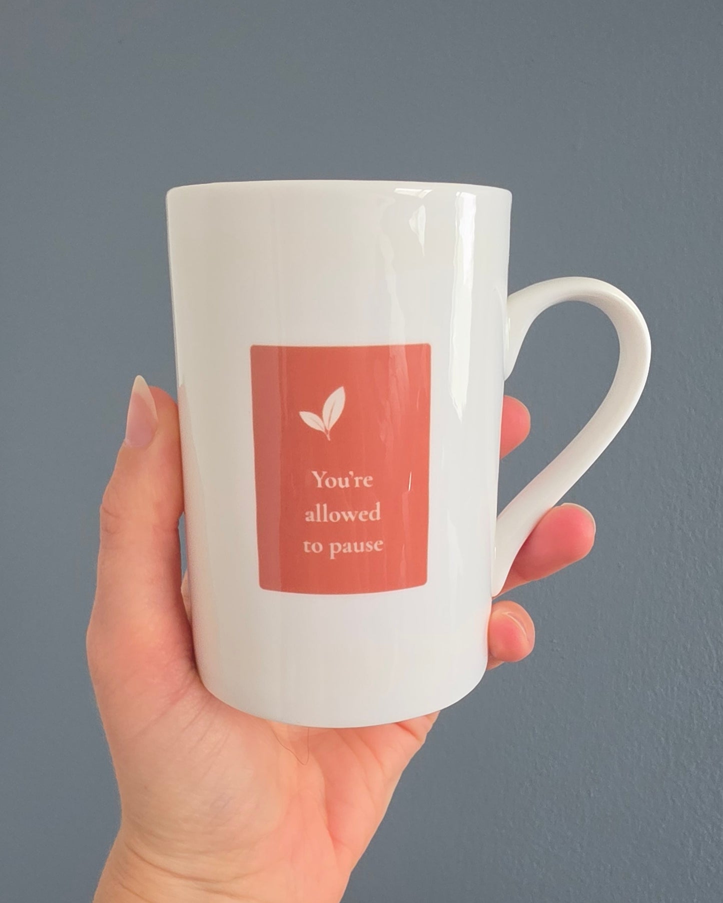 A hand holding a white ceramic mug with a terracota square graphic that reads 'You're allowed to pause' with a small leaf icon, set against a solid blue background.