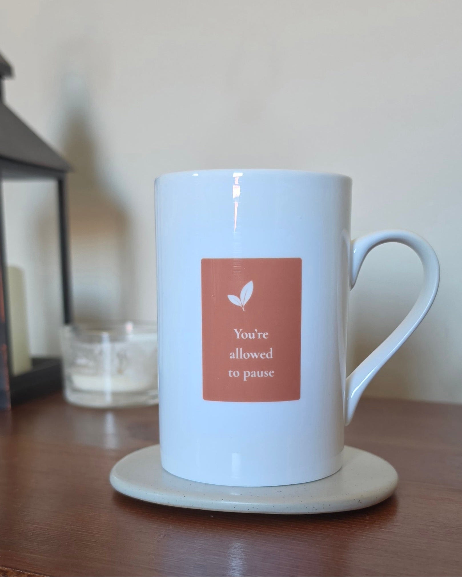 A white ceramic mug with a terracotta-colored square label that reads 'You're allowed to pause' with a small leaf icon, sitting on a beige coaster on a wooden table.