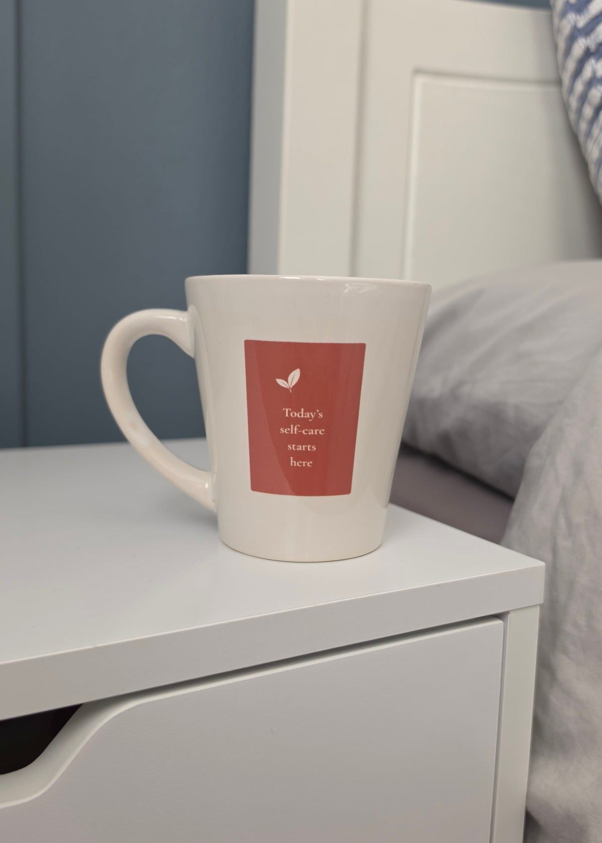 A white ceramic mug sitting on a white bedside table next to a bed with blue paneled walls.