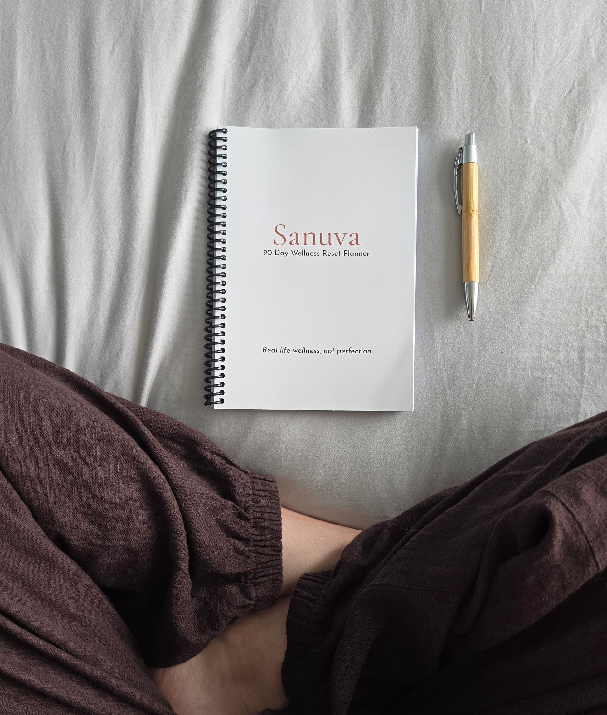 A white spiral-bound notebook with the brand name 'Sanuva' printed on the cover lies on a light gray fabric surface, flanked by a wooden pen on the right and crossed legs in dark brown pants in the foreground.