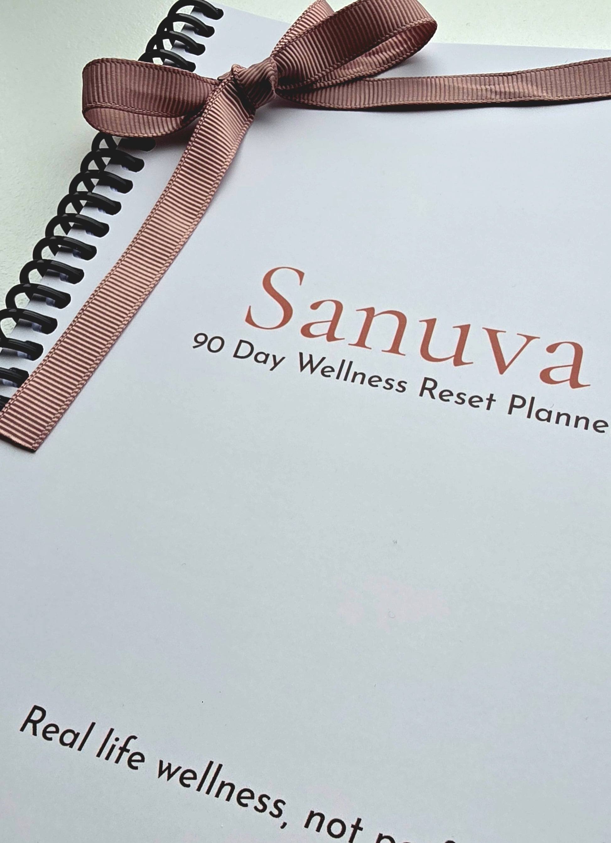 A close-up of a spiral-bound planner or journal cover with a brown ribbon tied in a bow across the top. The cover is white with the brand name 'Sanuva' in a muted reddish-brown font and the subtitle '90 Day Wellness Reset Planner' in smaller dark brown text.
