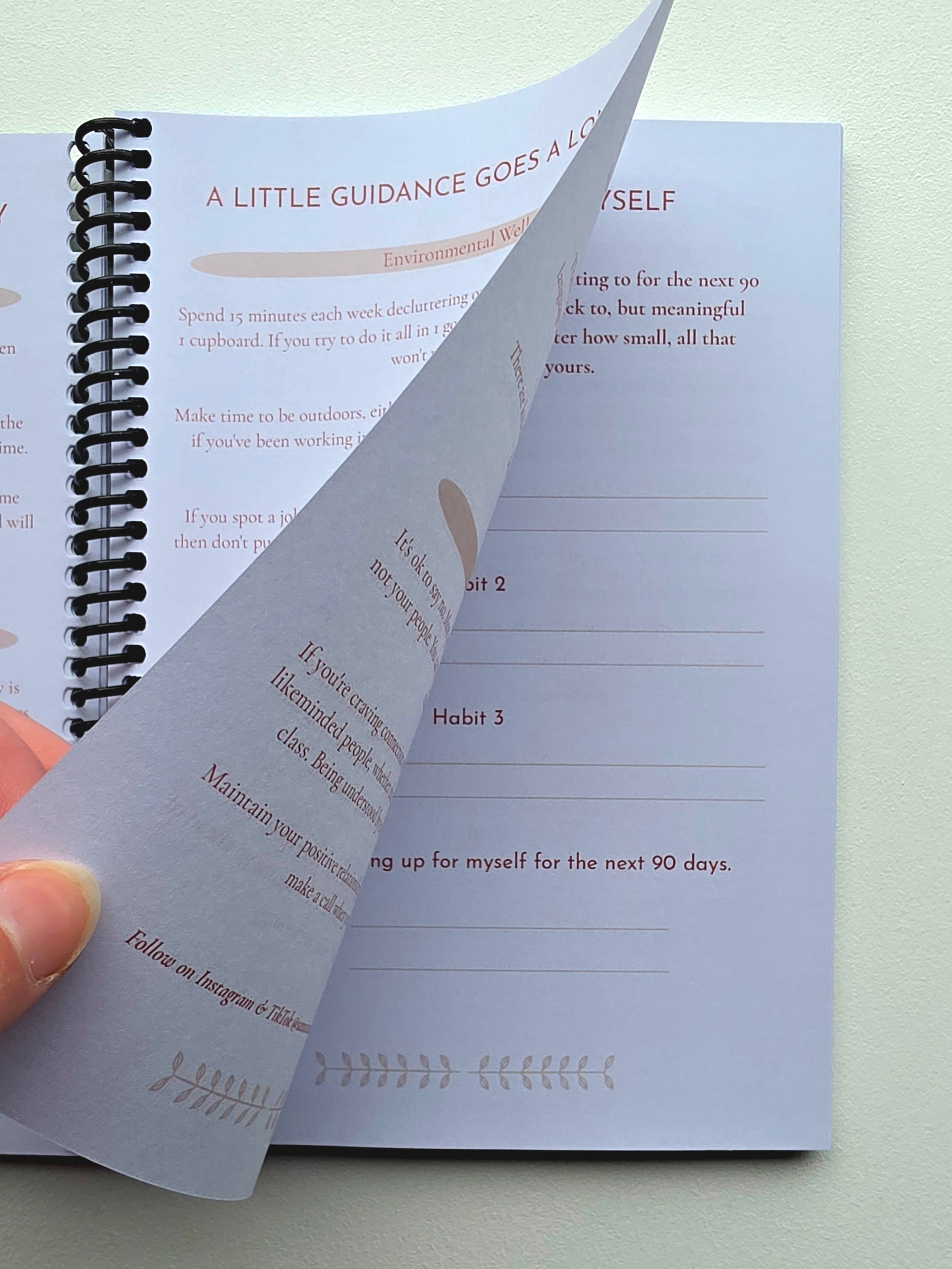 A spiral-bound journal or notebook shown open on a light surface, with a hand turning a page. The pages are white with soft text and decorative line illustrations