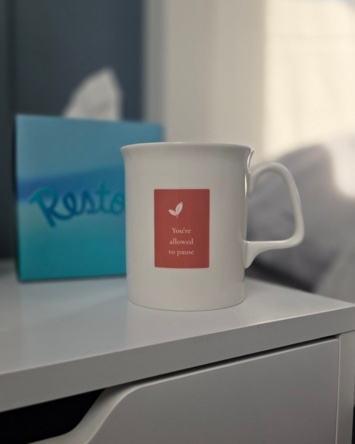 A white ceramic mug with a square label on the front, placed on a white bedside table next to a blue tissue box in a softly lit bedroom setting.