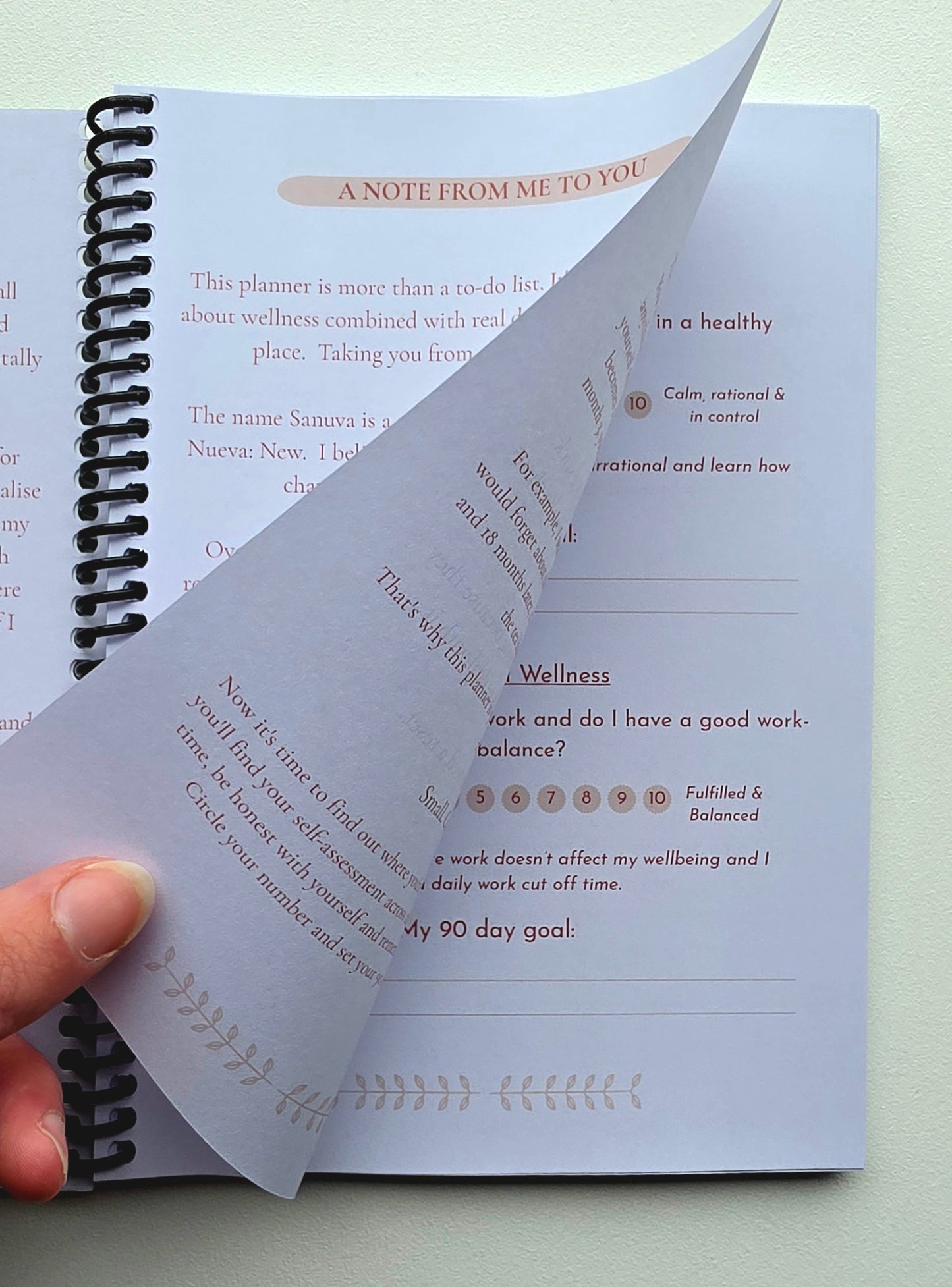 A close-up of a spiral-bound booklet or journal page being held open by a person's fingers. The page shows printed wellness prompts and text, with one sheet partially curled back against another open page on a light neutral background.