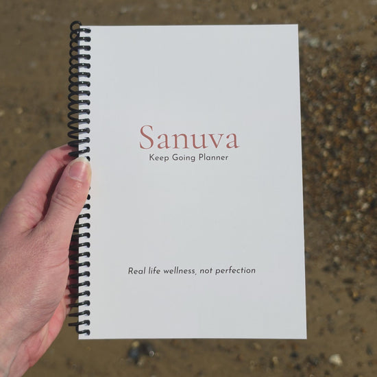 Video showing someones hand holding a planner which they turn over to show each side, while the sea tide flows back and forth in the background