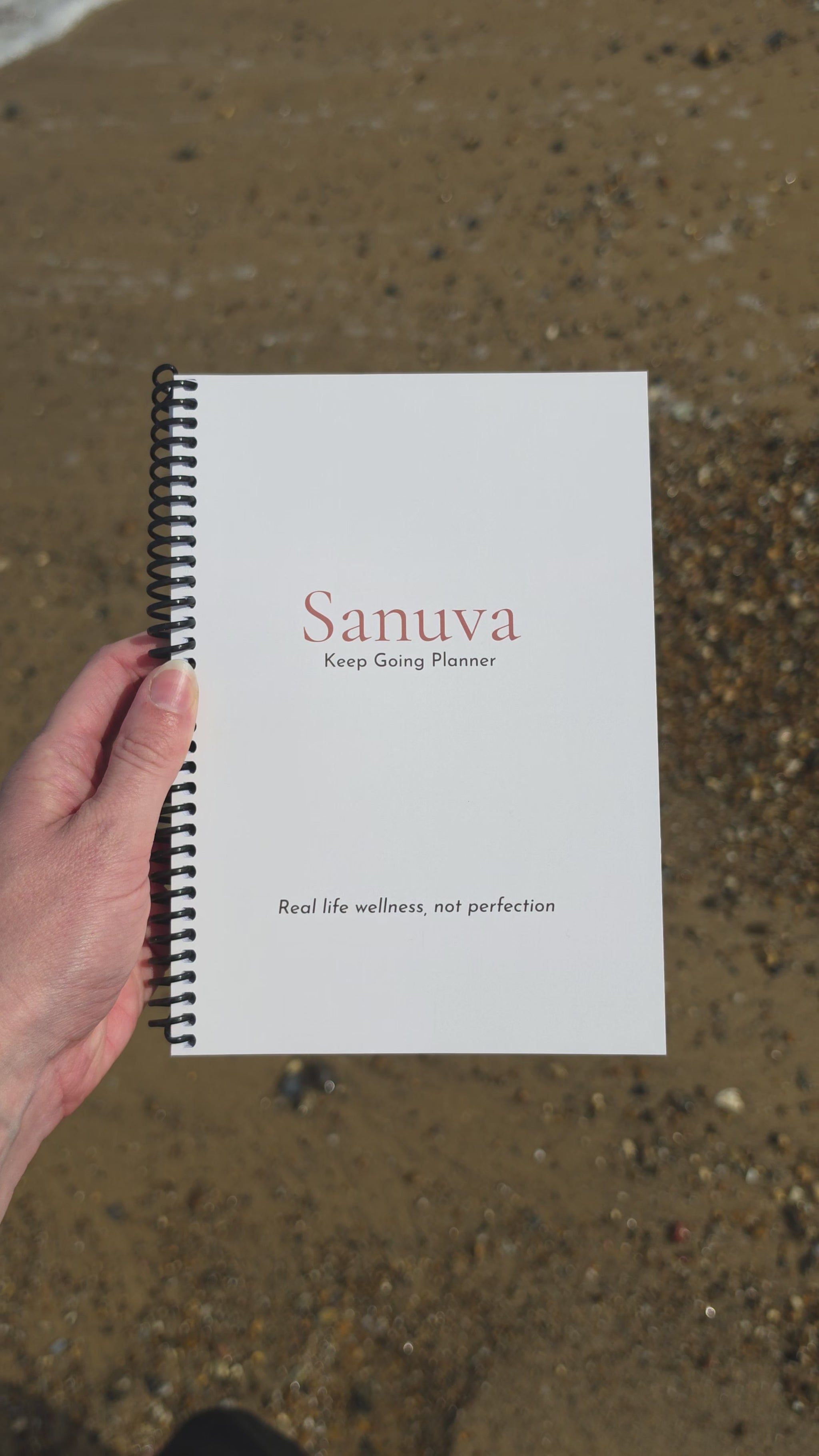 Video showing someones hand holding a planner which they turn over to show each side, while the sea tide flows back and forth in the background