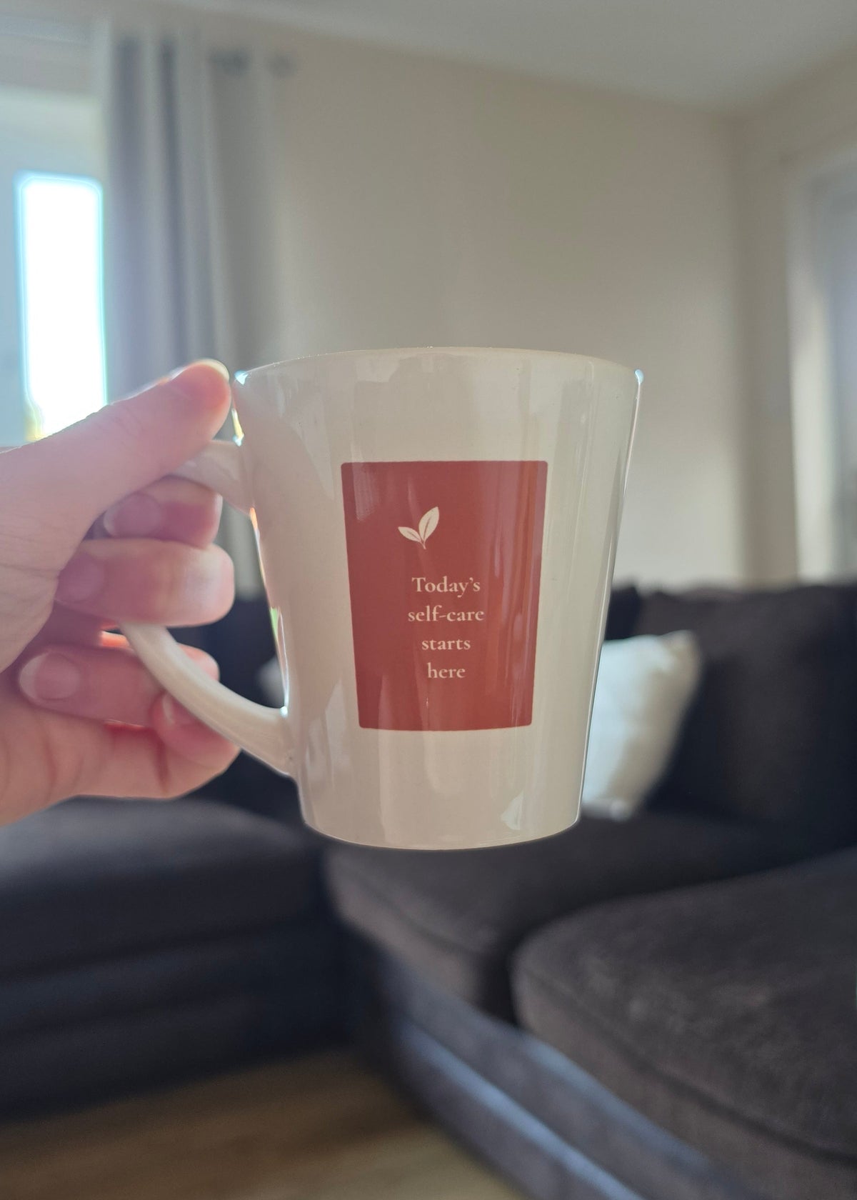 A hand holds a white ceramic coffee mug with a terracotta square label that reads 'Today's self-care starts here' with a small leaf graphic. The background shows a blurred living room with a dark sofa and light wood flooring.