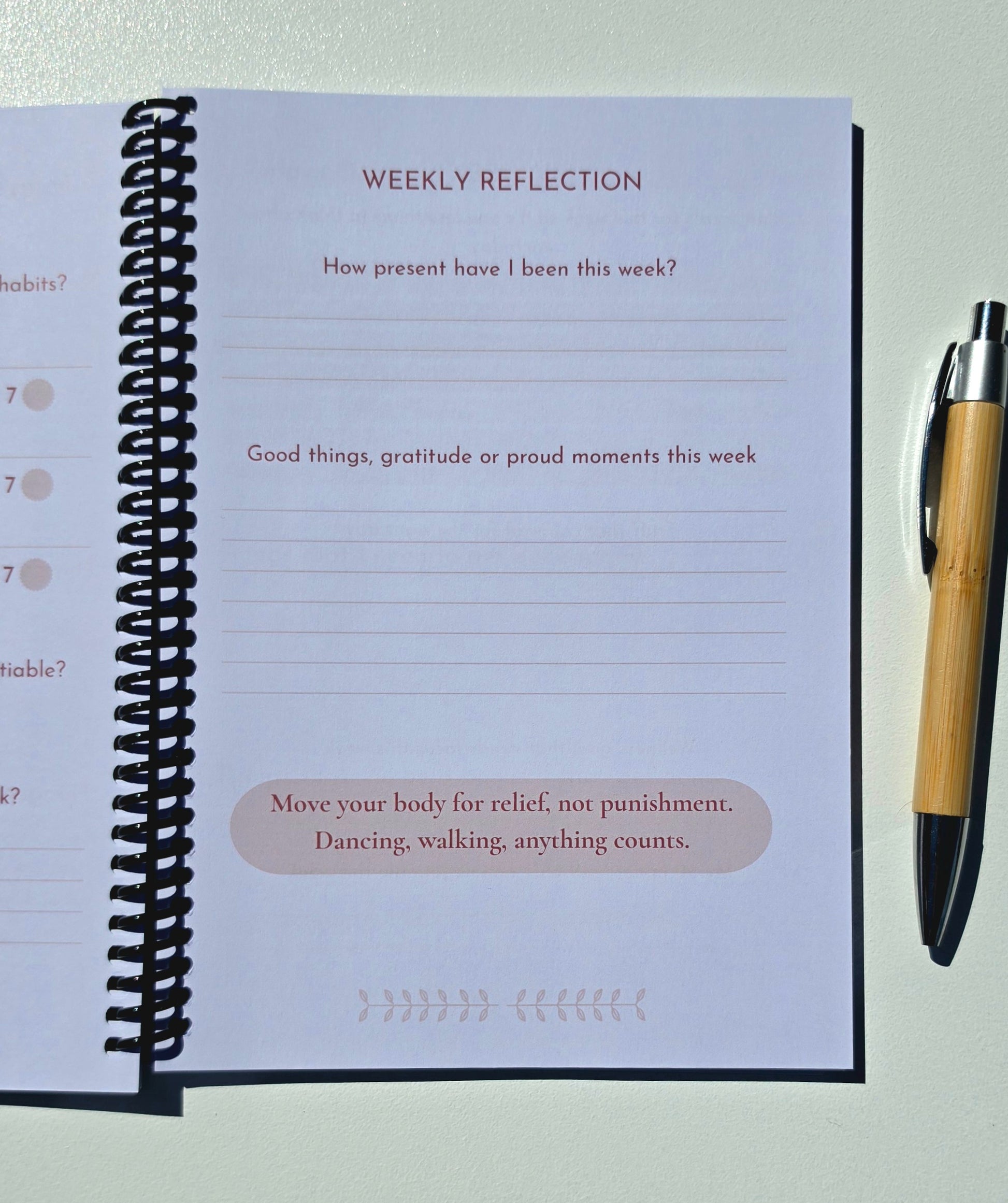 An open spiral-bound journal showing a weekly reflection page with lined writing areas and prompts, photographed on a light surface next to a black-and-wood pen.
